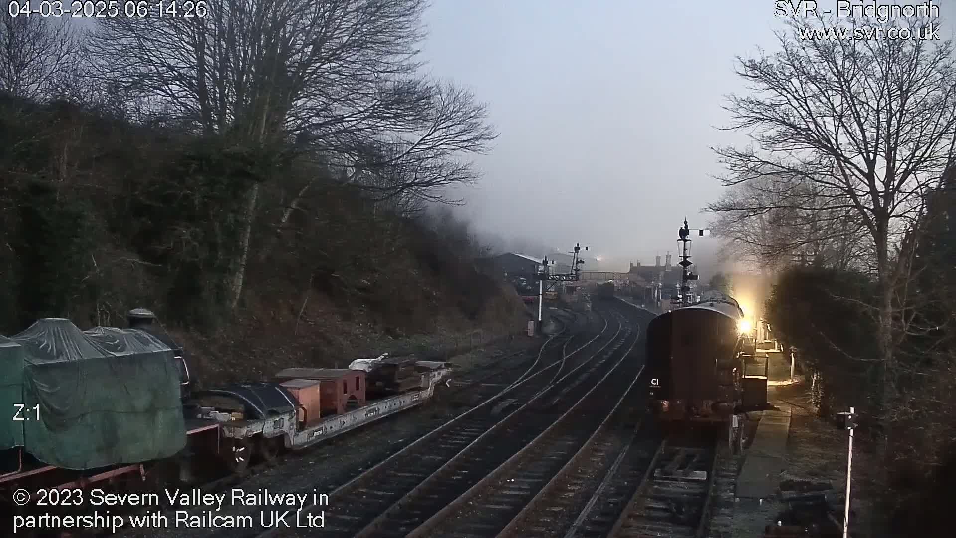 A steam train approaches a railway station on a foggy morning, passing by a covered railway car and other freight cars.