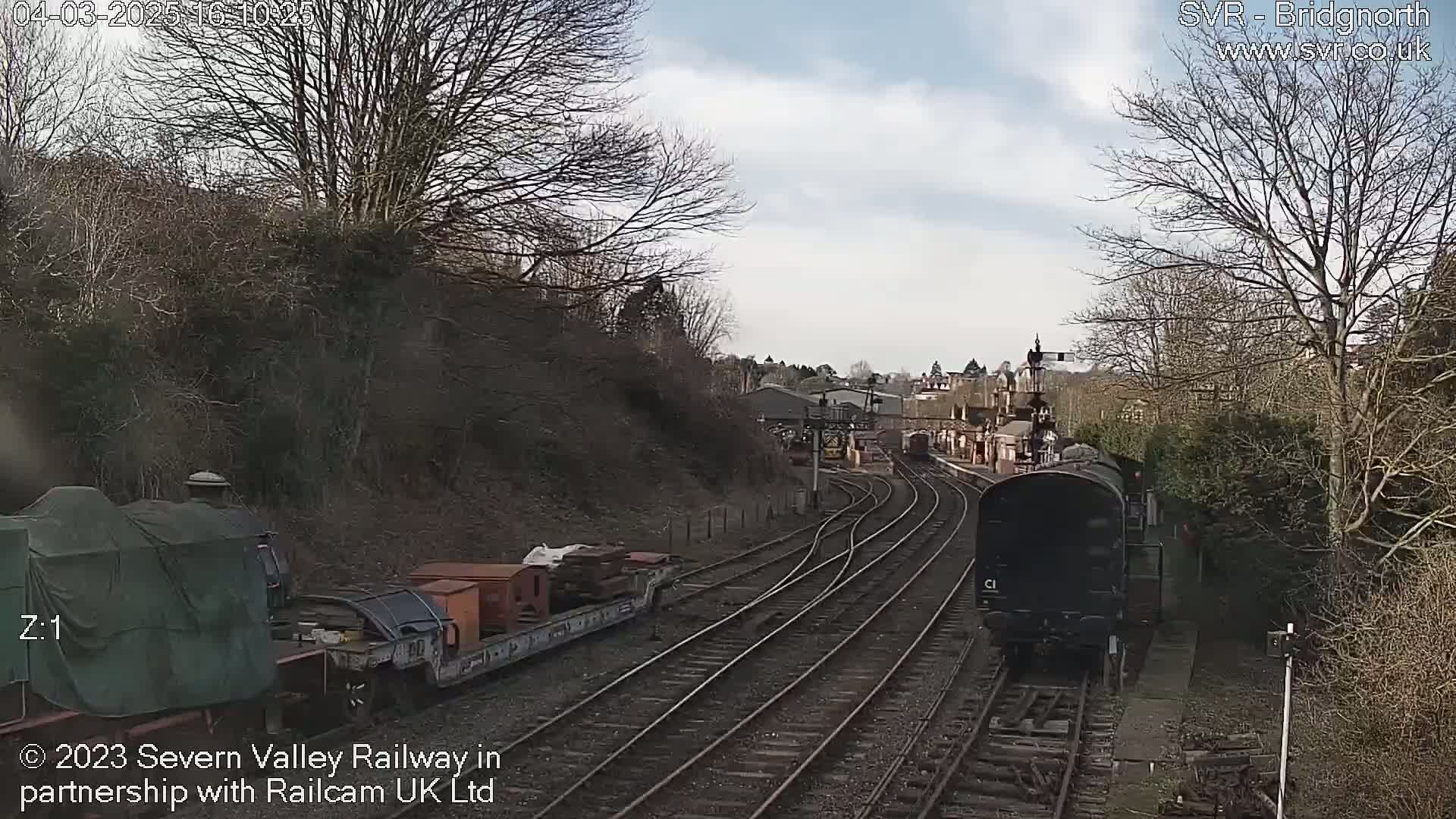 A train moves along railway tracks on a partly sunny day past a train station and bare trees.