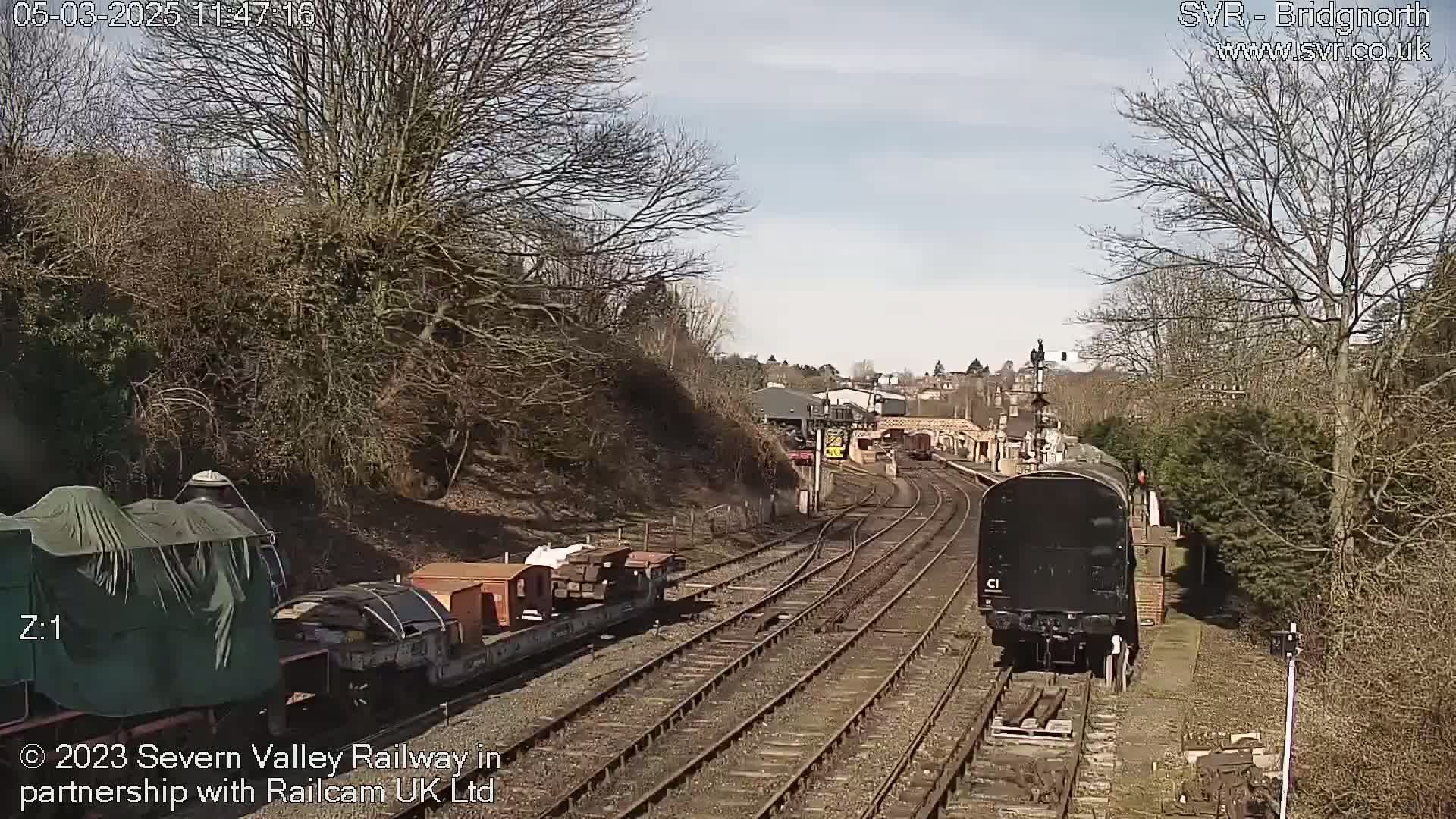 A black train is approaching a railway station on a sunny day, passing by a freight train carrying several orange cargo containers.