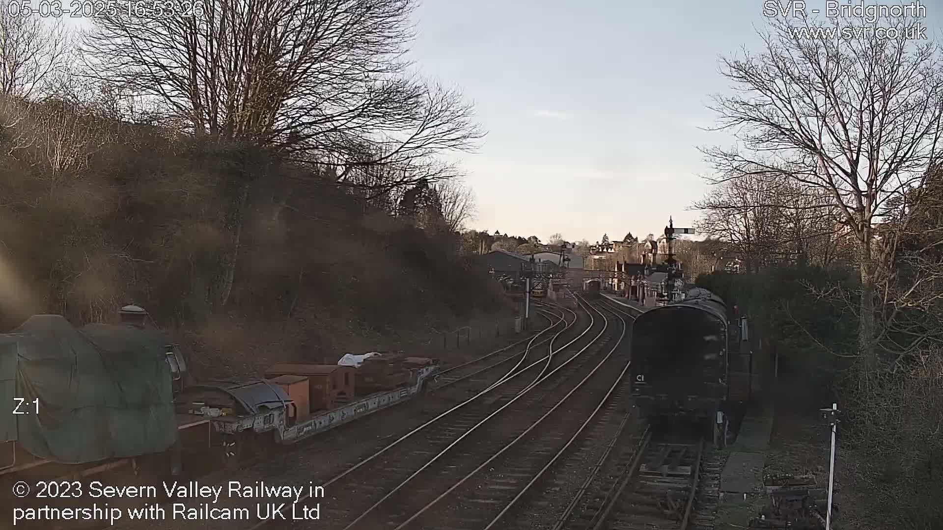 A dark-colored train approaches a railway station under a partly cloudy sky, with bare trees lining the tracks and hillsides.