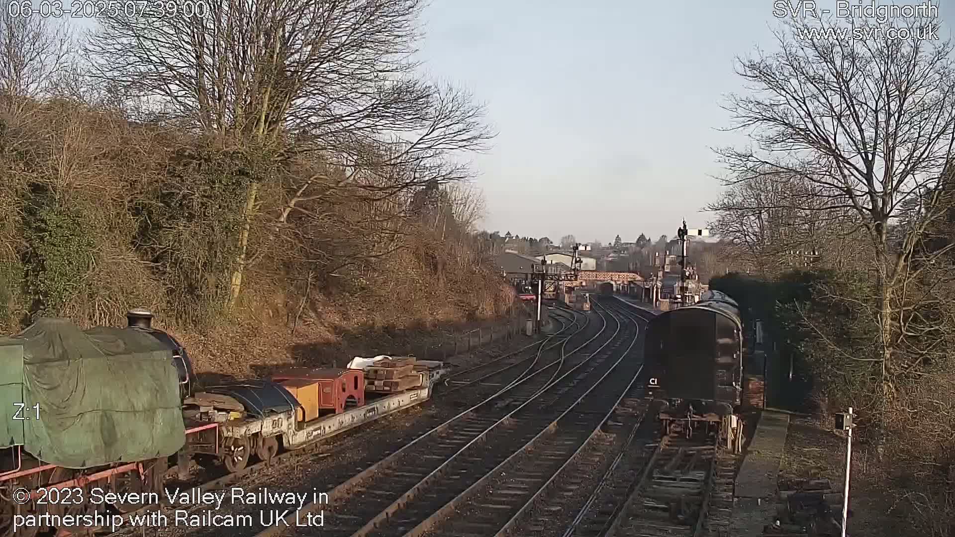 A sunny day at a railway station shows two trains on separate tracks, one covered and carrying cargo, the other a single dark engine.