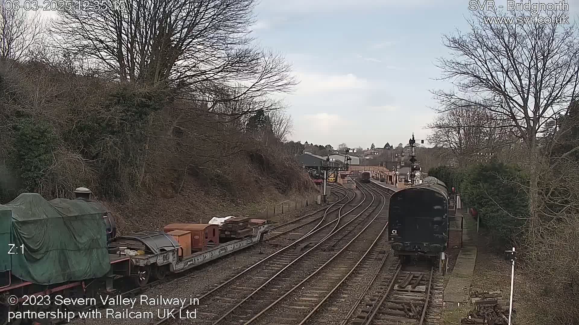 A train is moving along a railway track under a partly cloudy sky, passing by other railway cars and a hillside with bare trees.