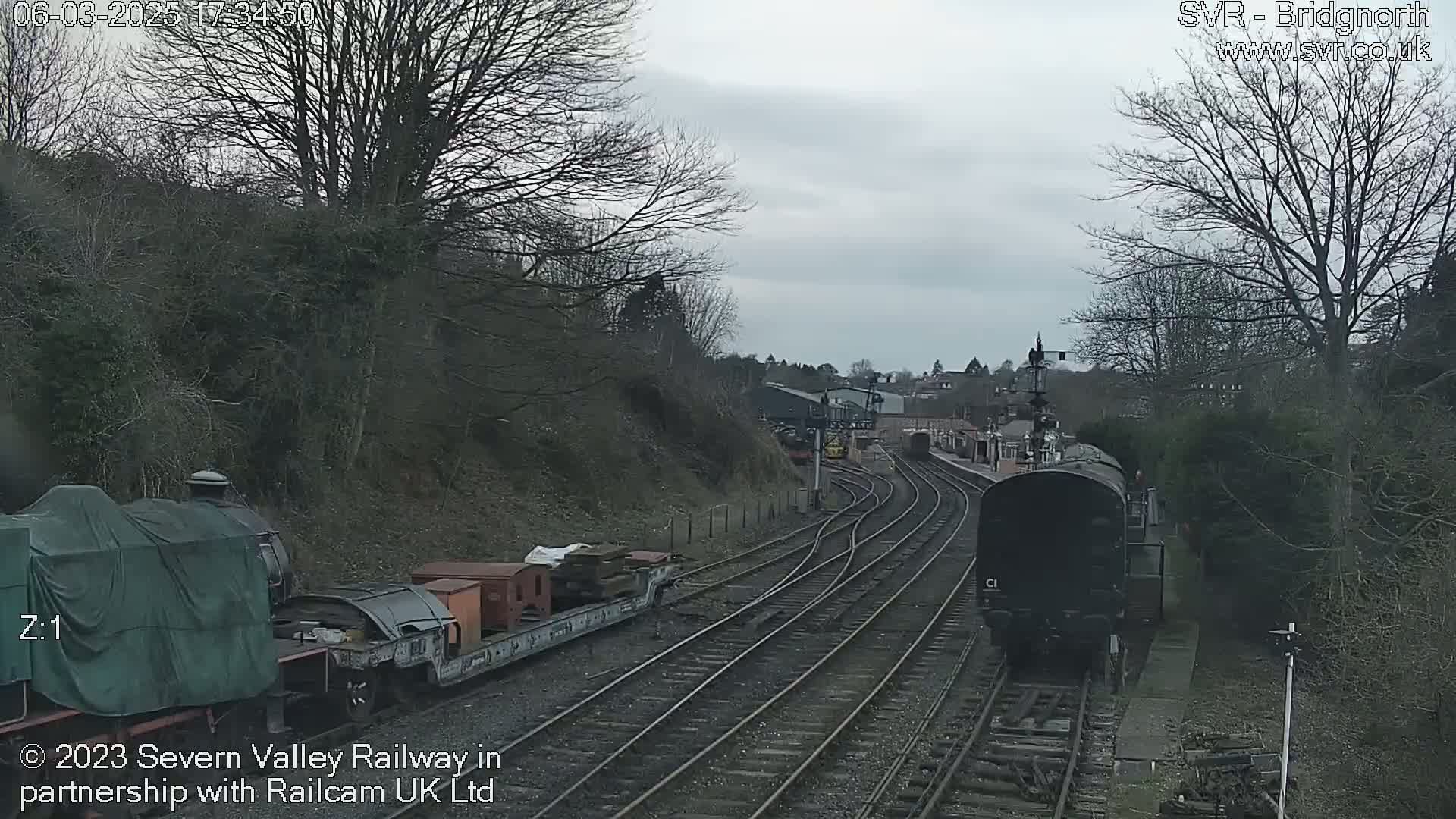 A dark-colored train travels along a track on an overcast day, passing another train partially covered by a tarp and some cargo cars.