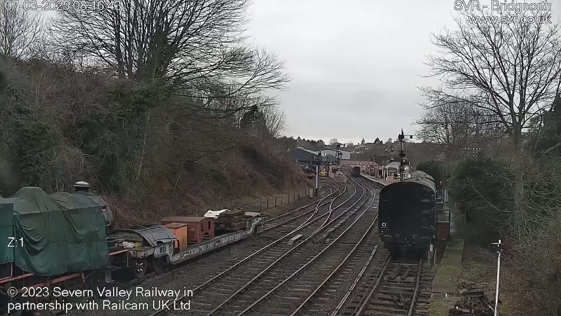 A train approaches a railway station on an overcast day, passing alongside a stationary train carrying cargo.