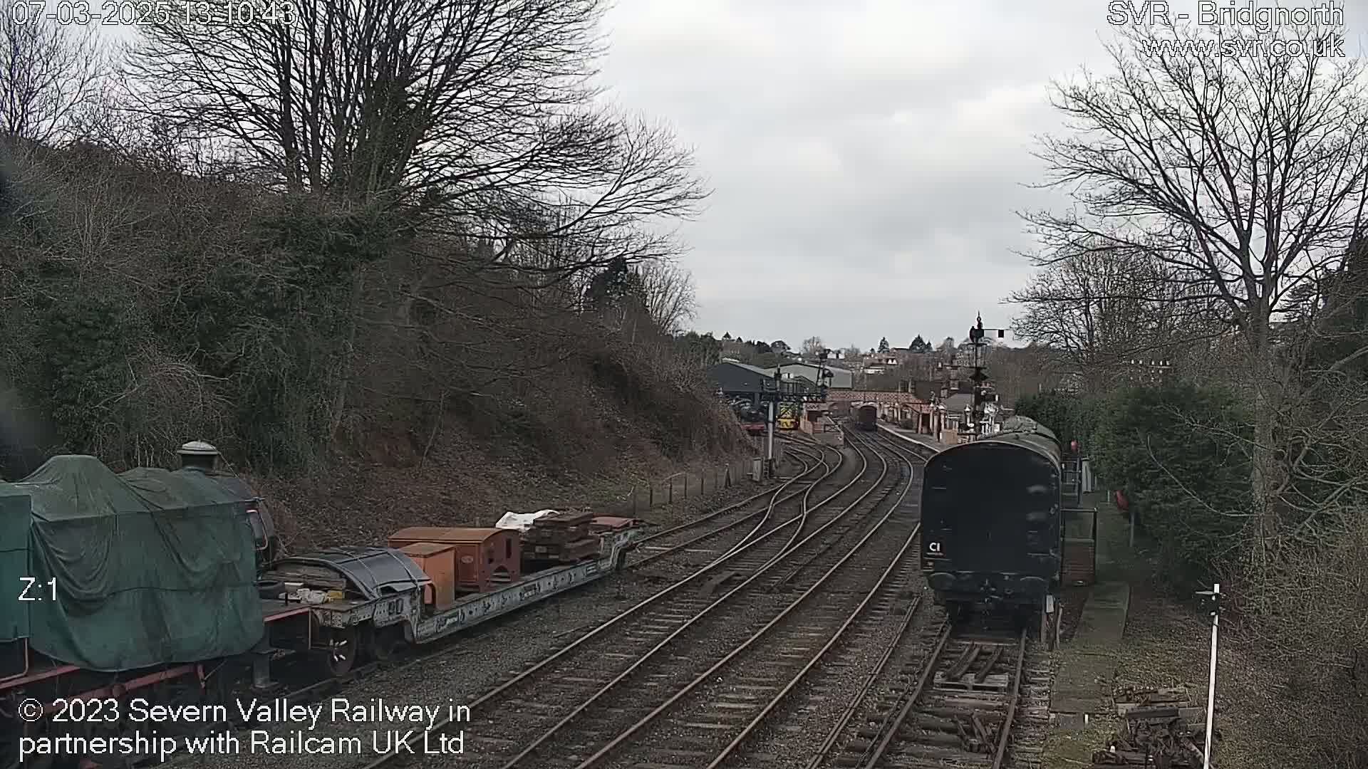 A train approaches a railway station on a cloudy day, passing another train carrying cargo.