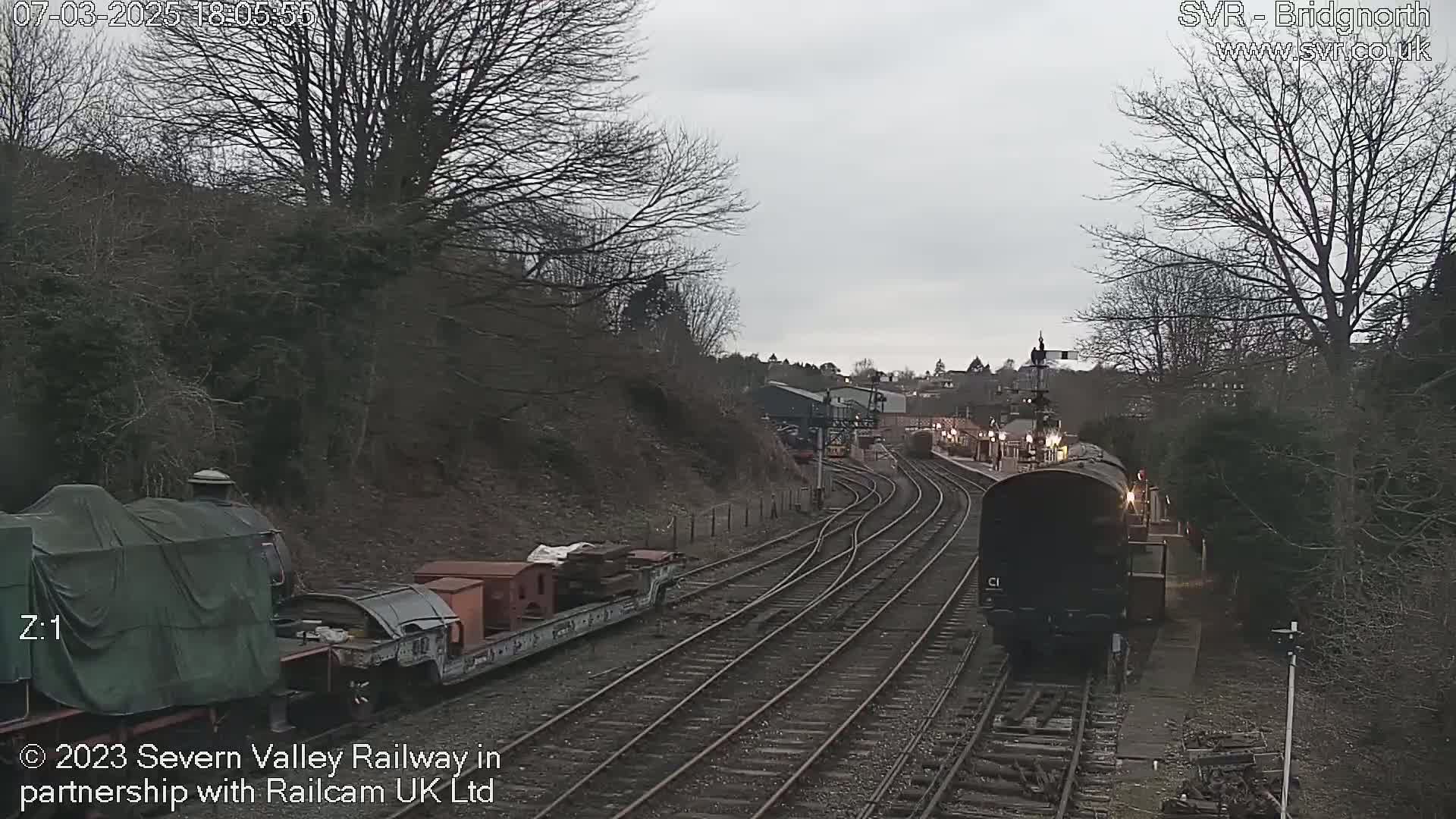 A train approaches a railway station on an overcast day, passing by a stationary train and railway cars.