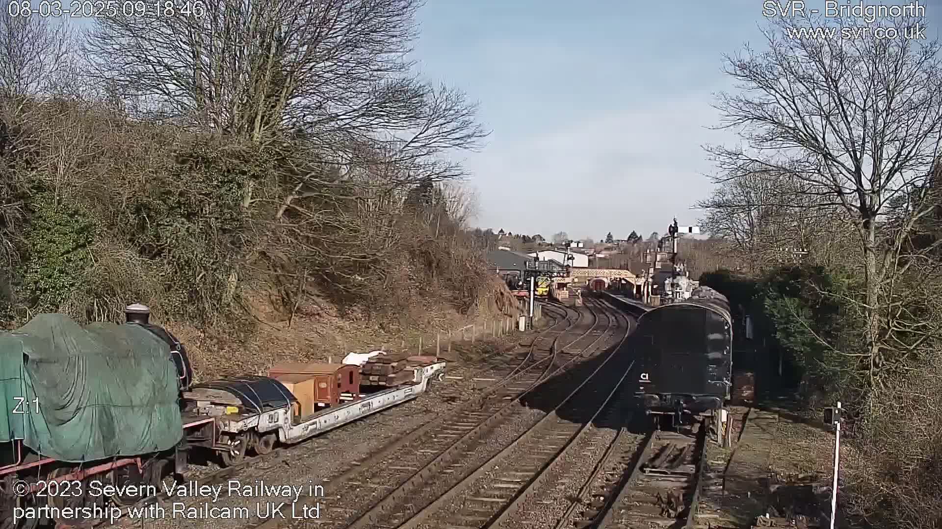 A sunny day at a train station with two trains on separate tracks, one covered and carrying cargo, the other a dark-colored passenger car.