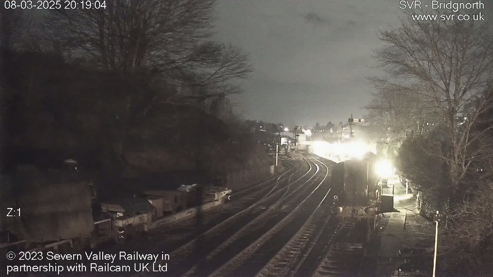 A nighttime view of a train station with a train approaching, under a dark, cloudy sky.