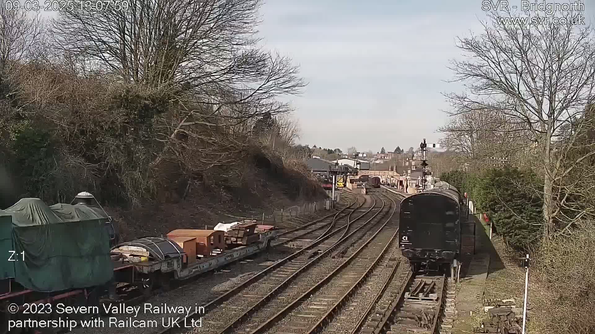 A sunny day at a railway station shows two trains on parallel tracks, one covered in a tarp and the other a black diesel locomotive.