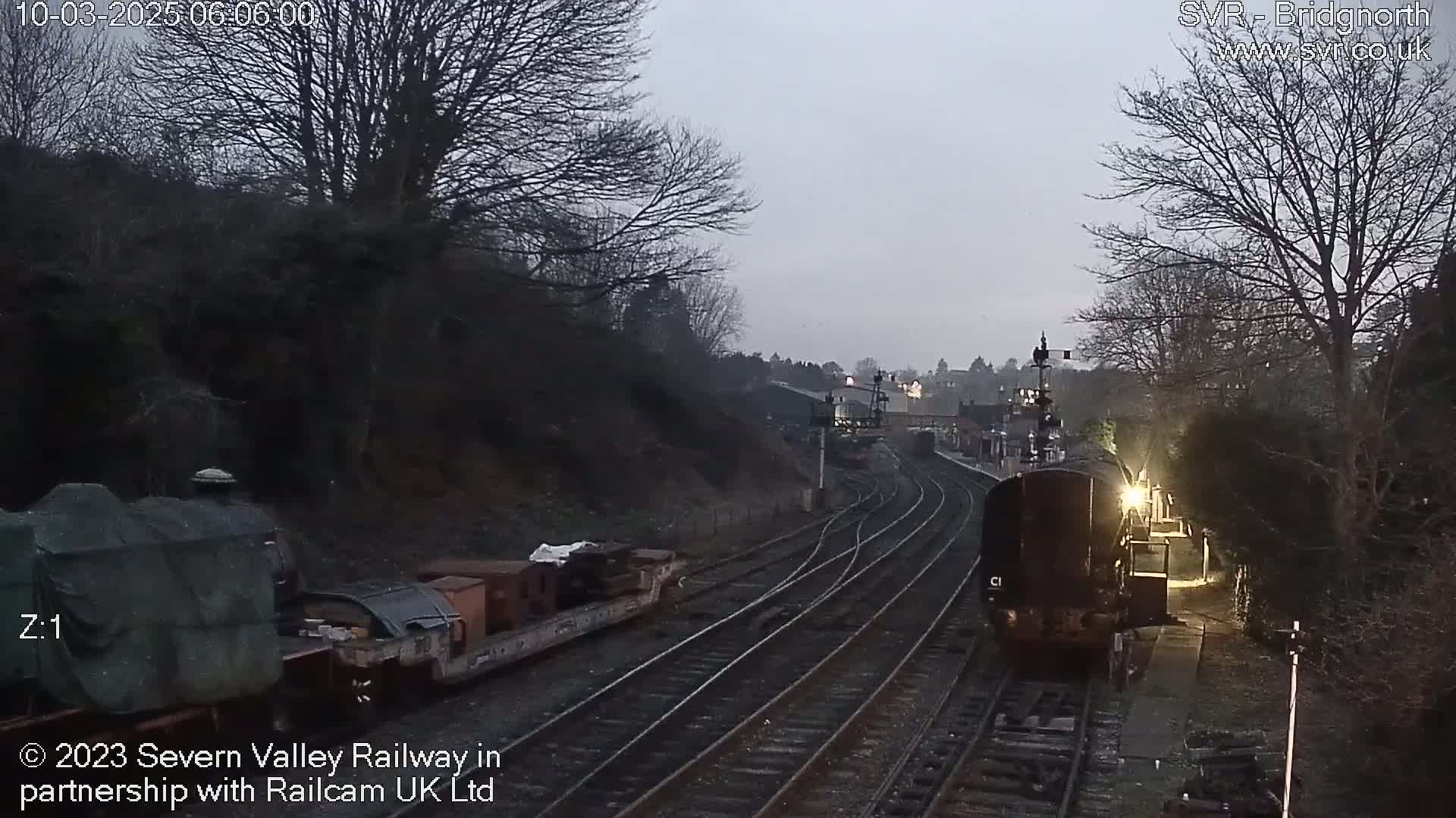 A diesel locomotive approaches a train yard on a cloudy day, passing a stationary train car.