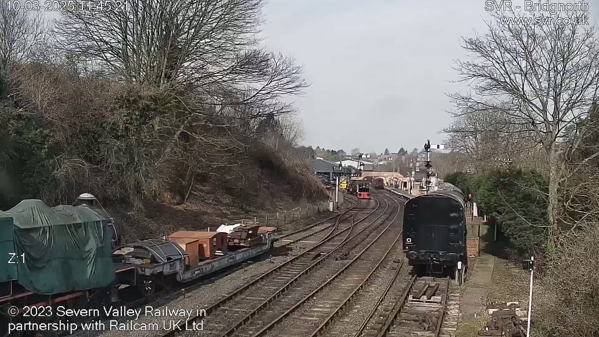 A train carrying freight cars moves along a railway track under a mostly cloudy sky, passing other trains and railway structures.