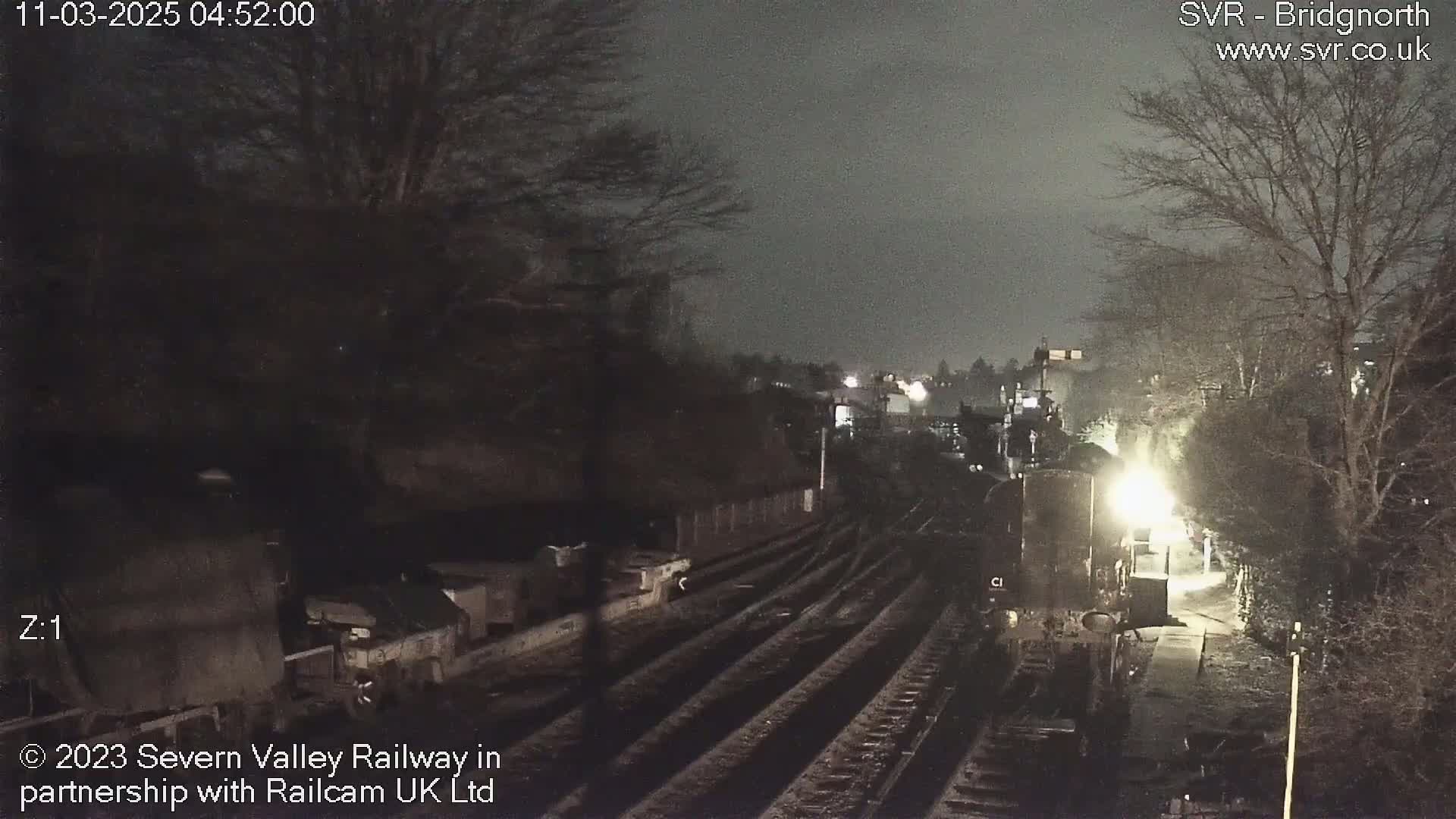 A nighttime view of a train station platform with train tracks extending into the distance, under a dark, overcast sky.