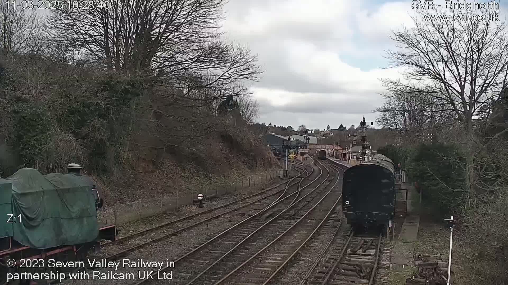 Two trains are visible on a railway track, one covered and stationary, the other dark-colored and moving, under a cloudy sky.
