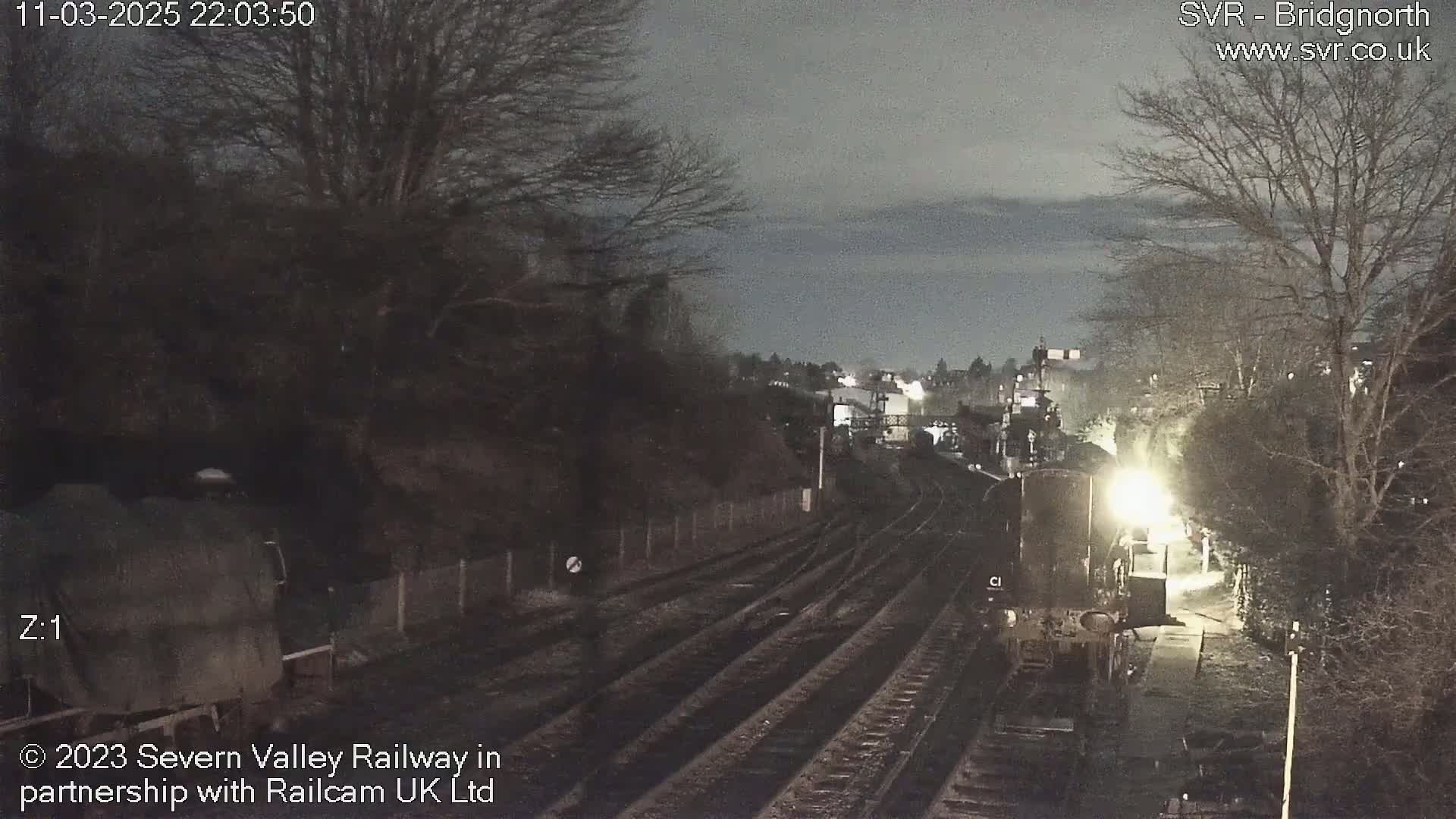 A nighttime view of a railway station with a train partially visible, under a dark, cloudy sky.