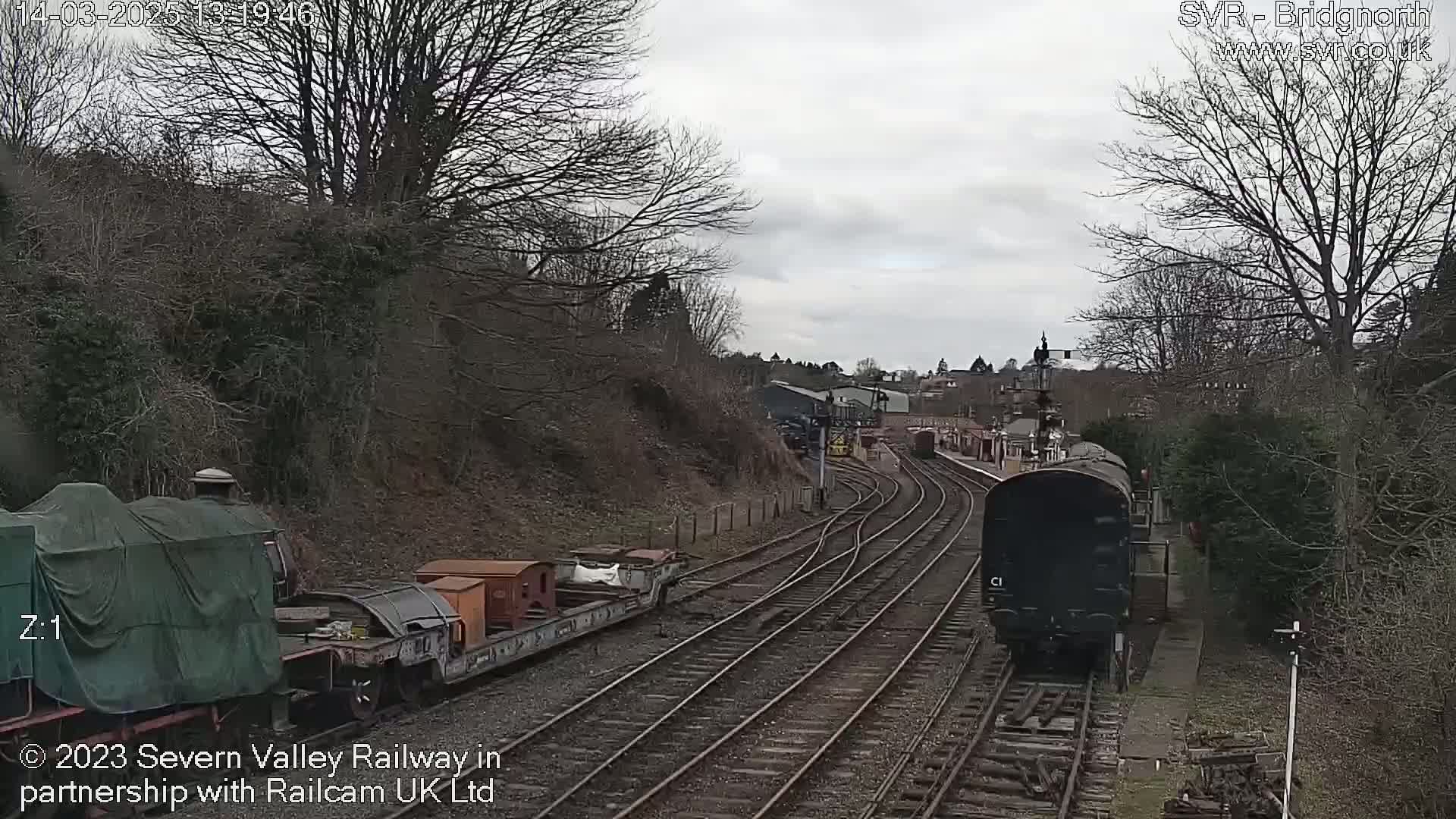 A dark-colored diesel locomotive approaches a railway station on an overcast day, passing several freight cars and a covered engine.