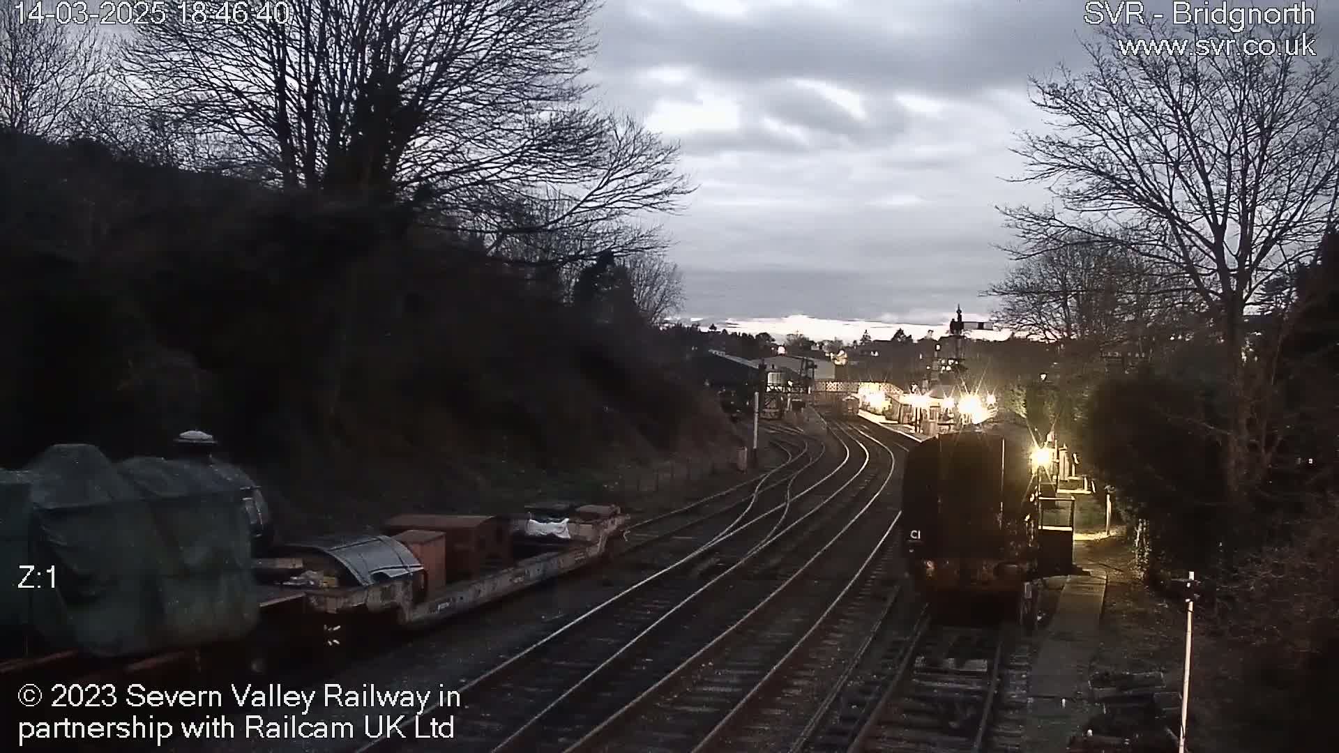 A train is approaching a station under a cloudy, dusky sky.