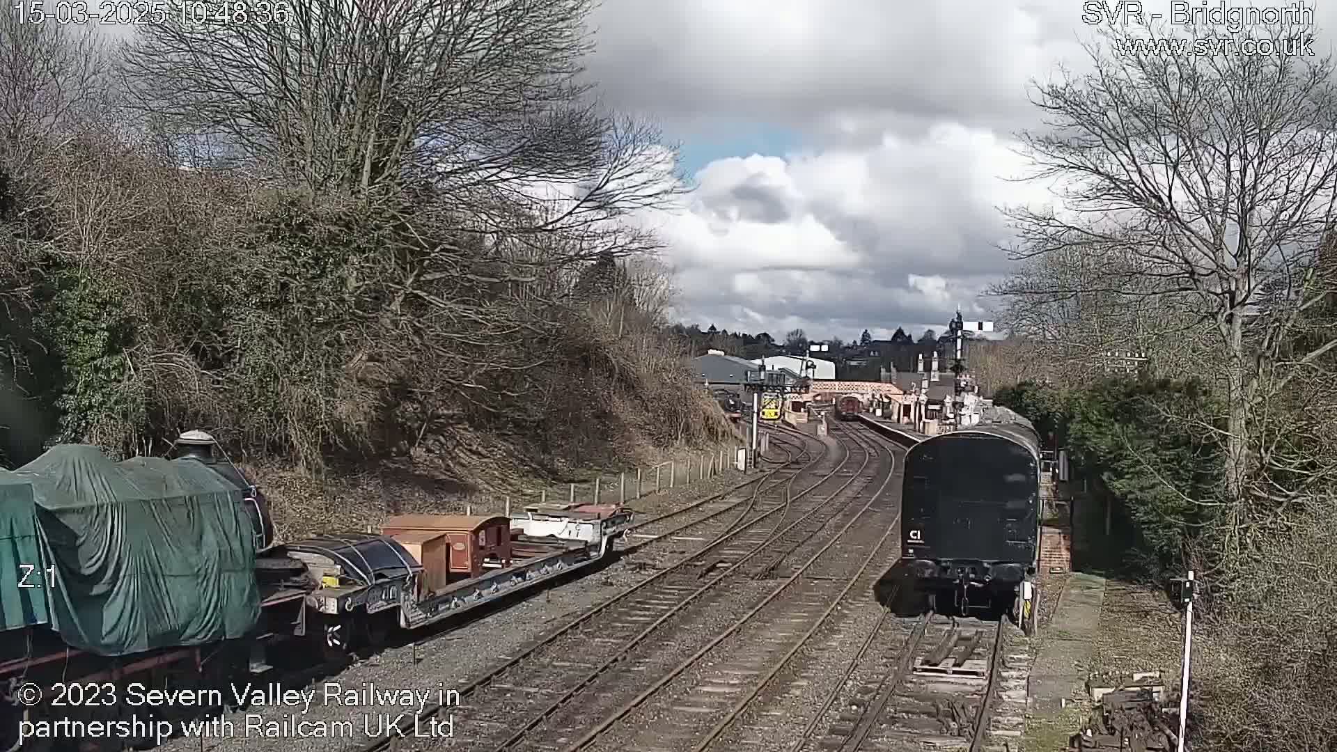 Two trains, one covered and one uncovered, are on a railway track under a partly cloudy sky.