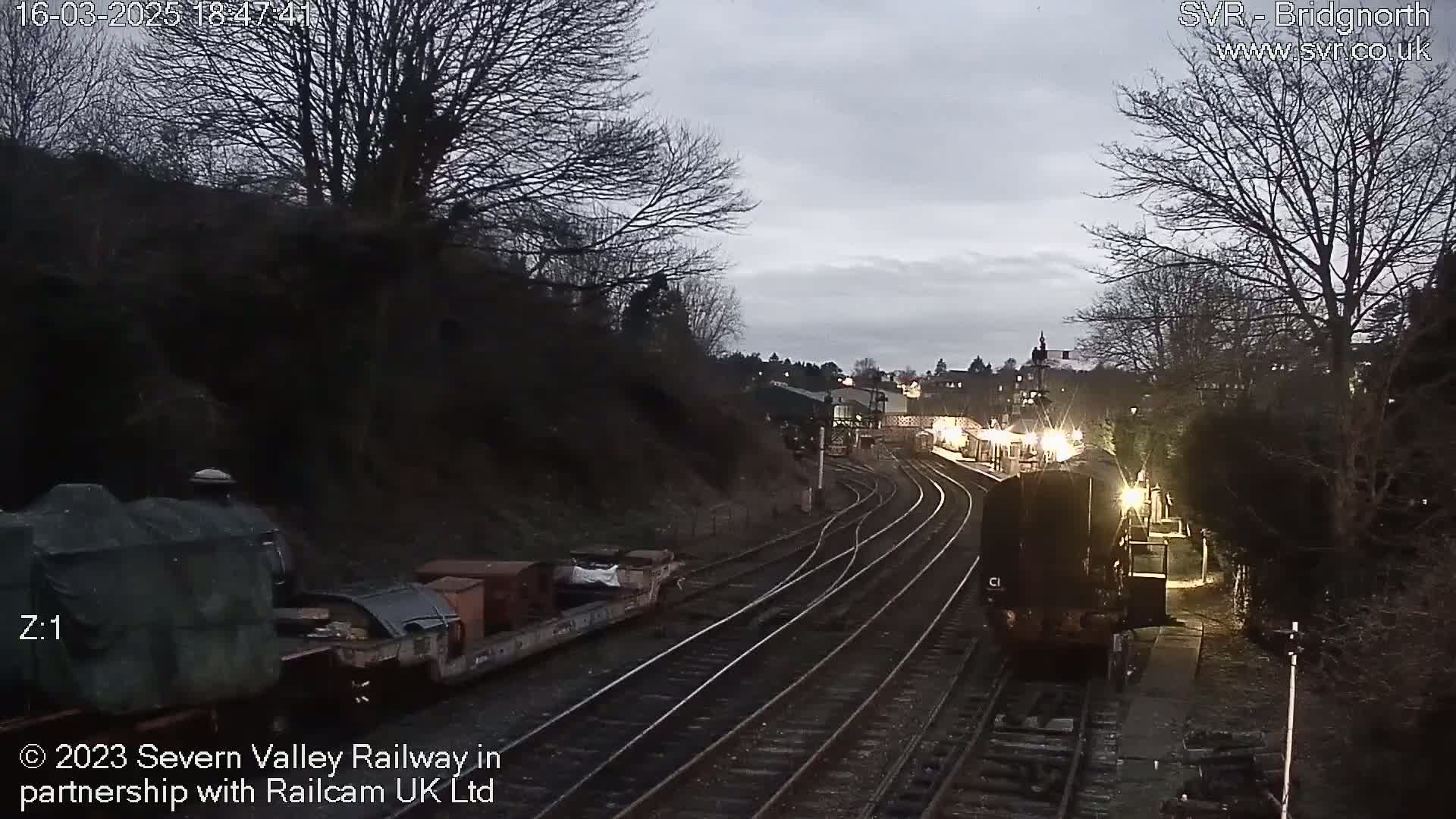 A train approaches a dimly lit railway station at twilight under a cloudy sky.