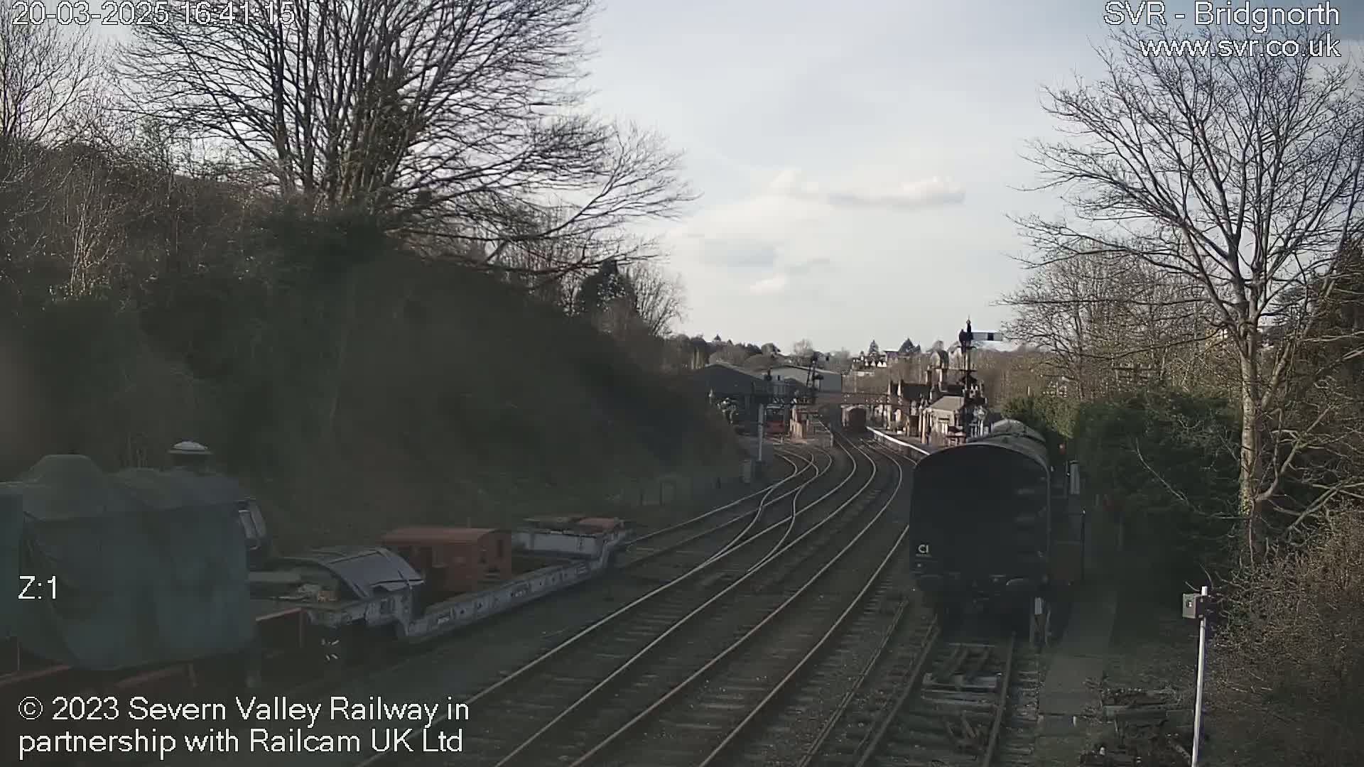 A dark-colored train approaches a railway station on a partly cloudy day, with bare trees lining the tracks.