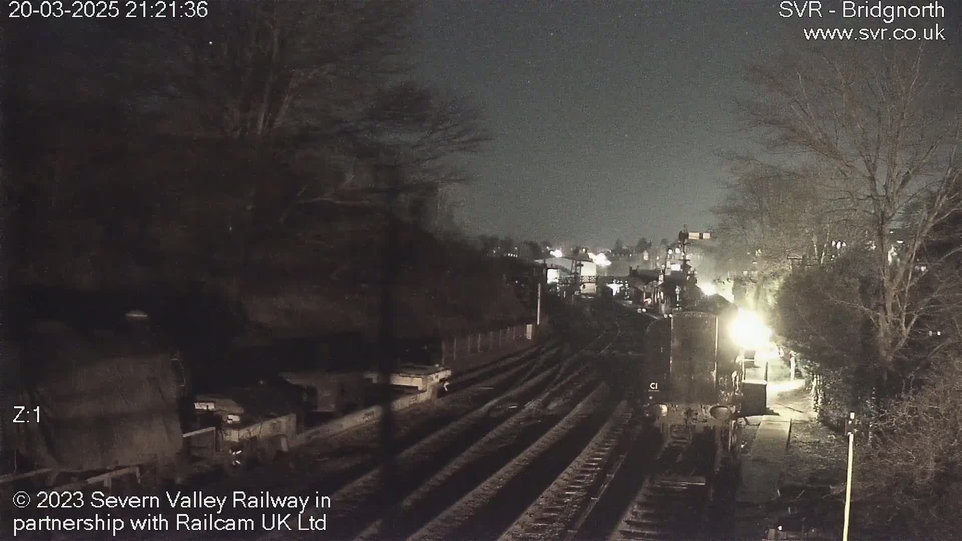 A nighttime view of a train station with a train partially visible on the tracks, under a dark sky.