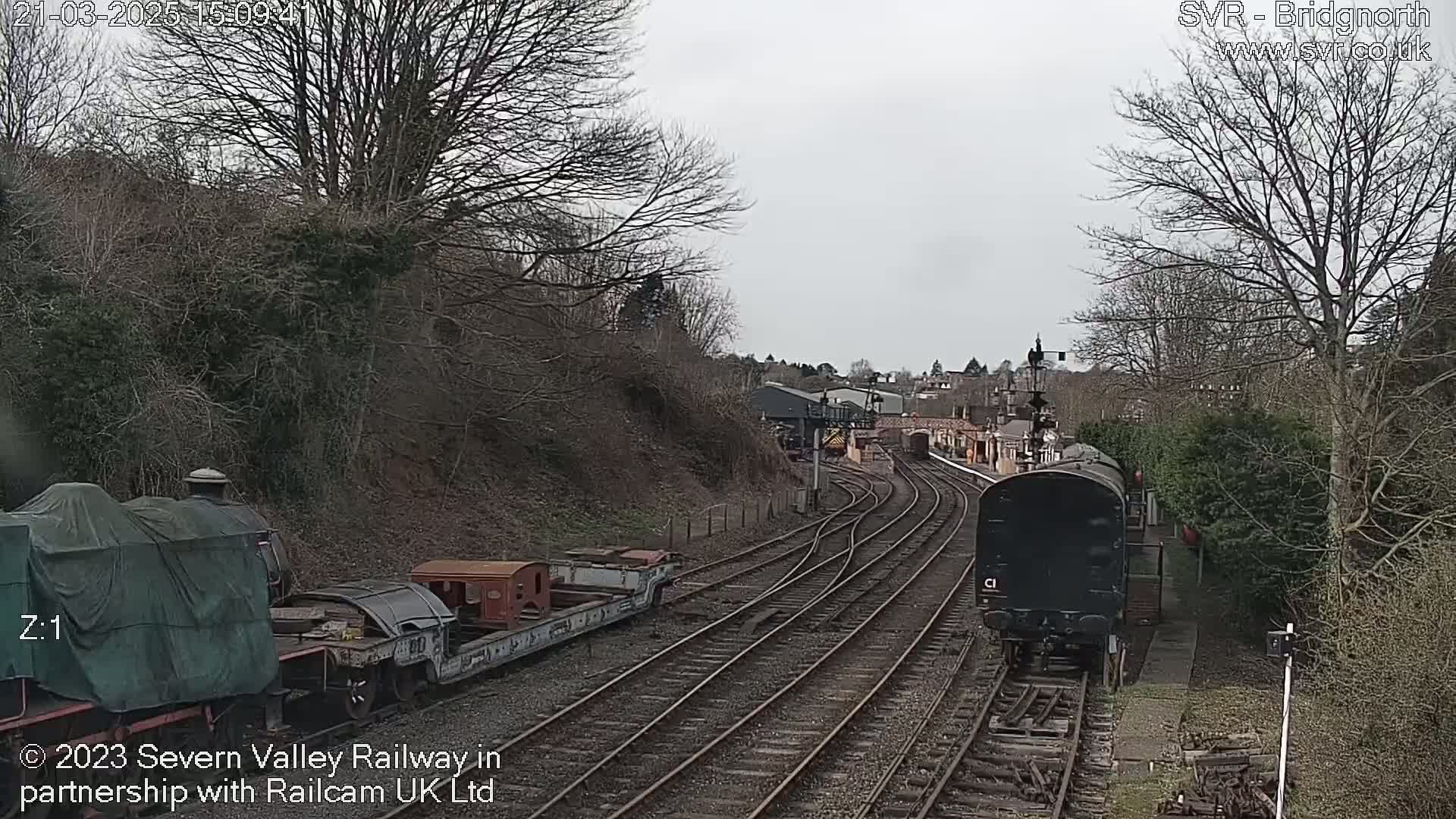 A dark-colored train moves along a railway track toward a station, passing by a covered train car and other railway infrastructure under an overcast sky.