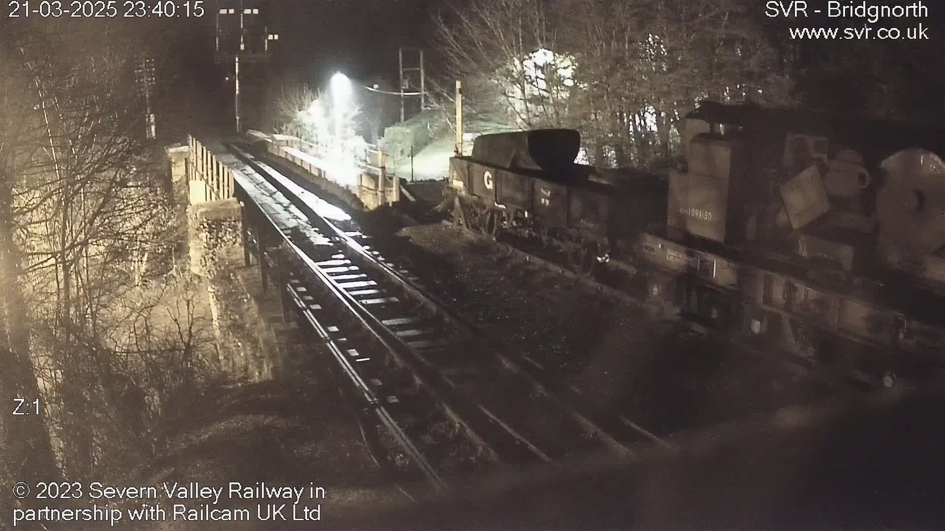 A dark-colored train sits on a set of tracks at night, partially visible beside a stone bridge.