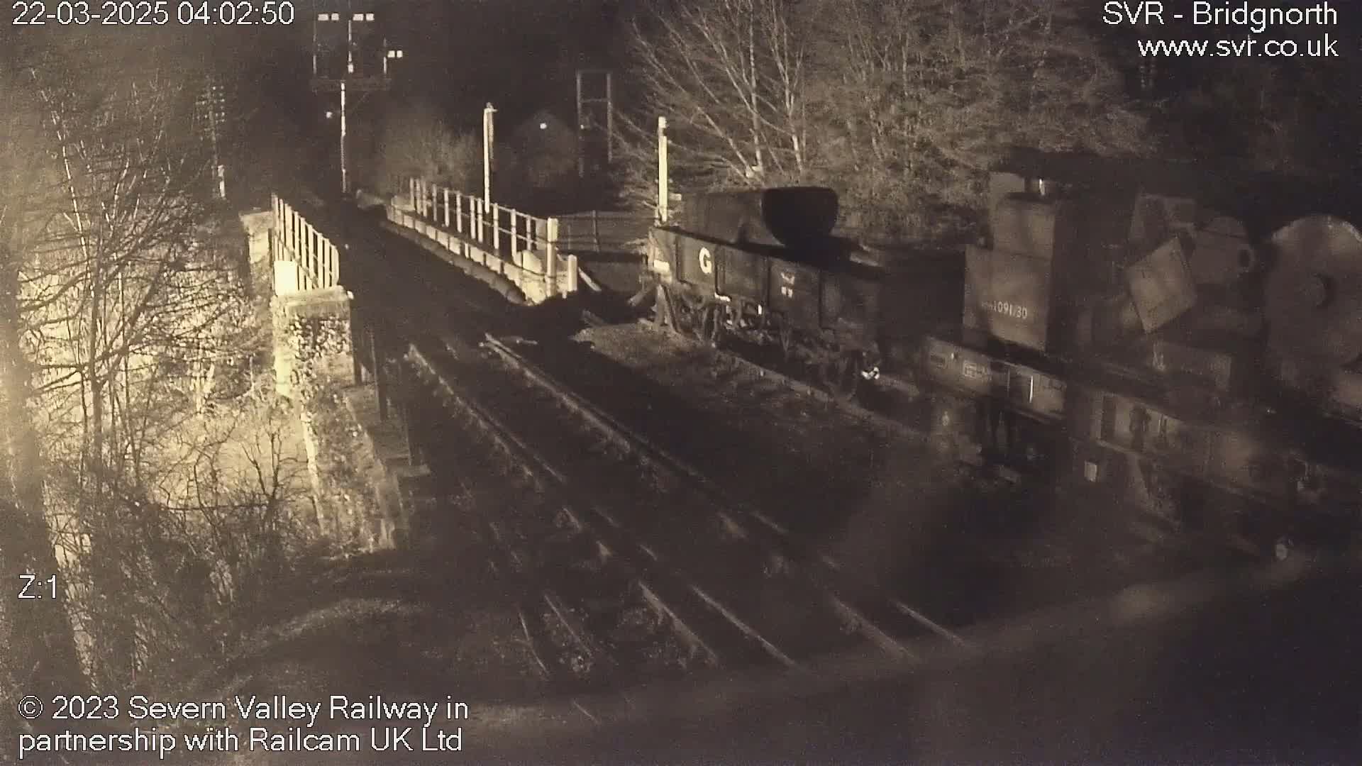At night, a freight train sits on railroad tracks beside a stone wall and trees.
