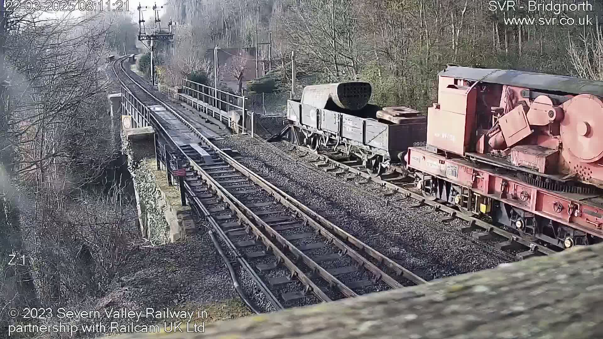 A red railway crane and a dark freight car are parked on a railway track next to a stone bridge on a sunny day.