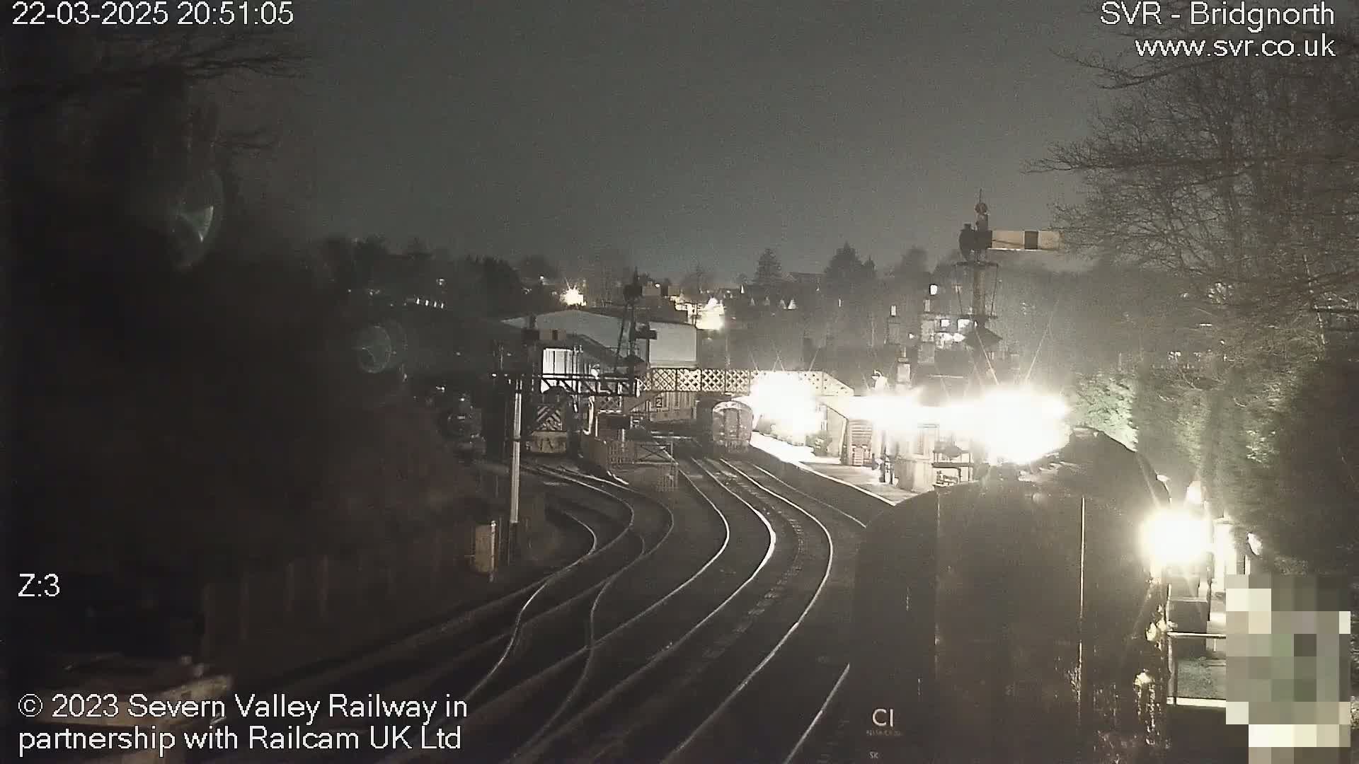 A nighttime view of a train station with several train tracks and a train partially visible, under a dark, overcast sky.