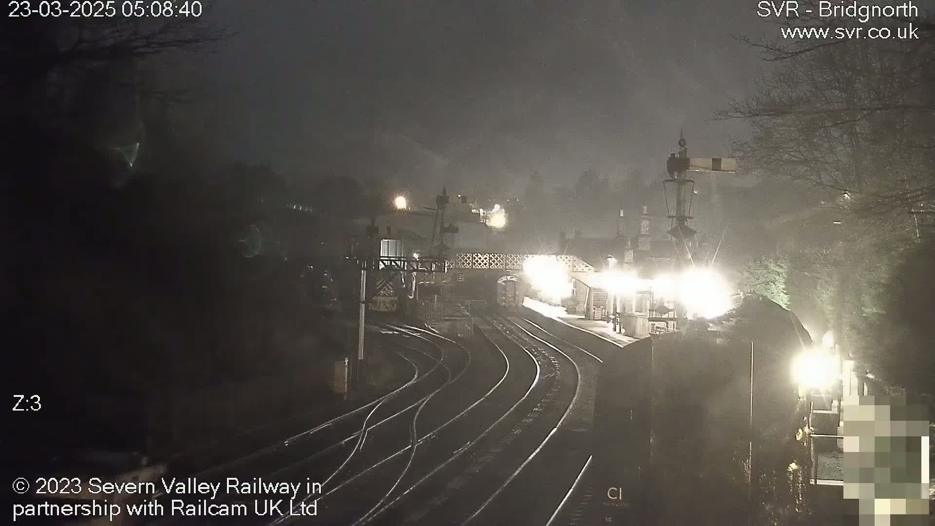 A nighttime view of a train station with several train tracks, under foggy or misty conditions.