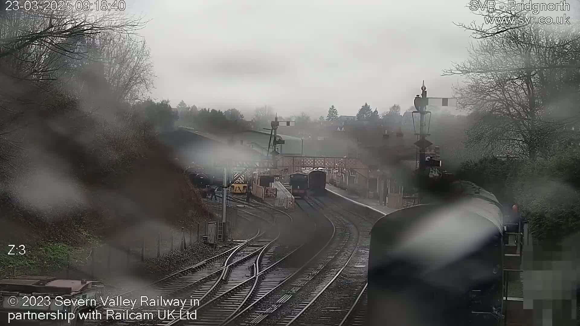A misty, overcast day at a train station shows several train cars on a track curving through the station.