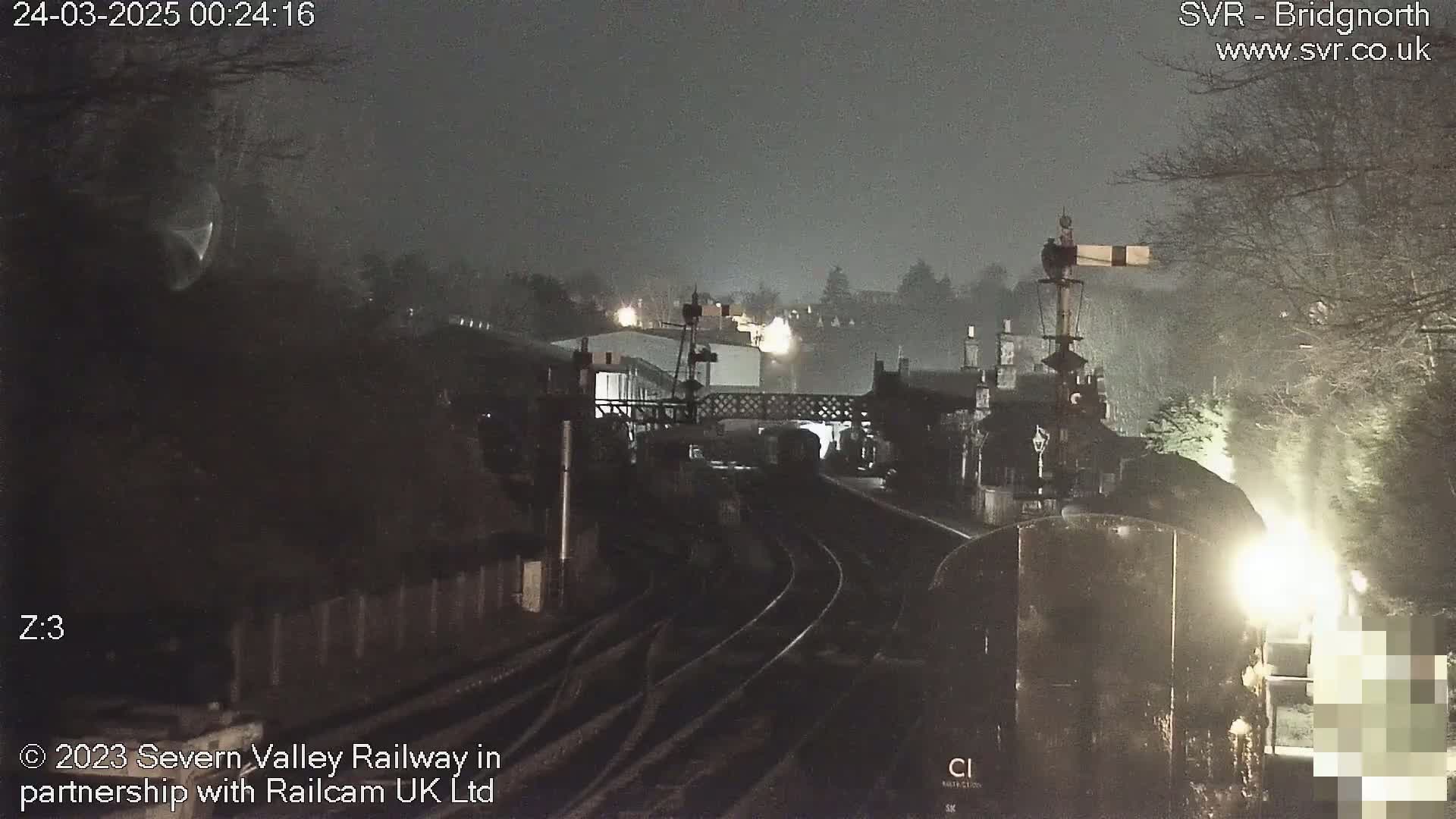 A nighttime view of a train on railway tracks near a station under dark, possibly overcast conditions.
