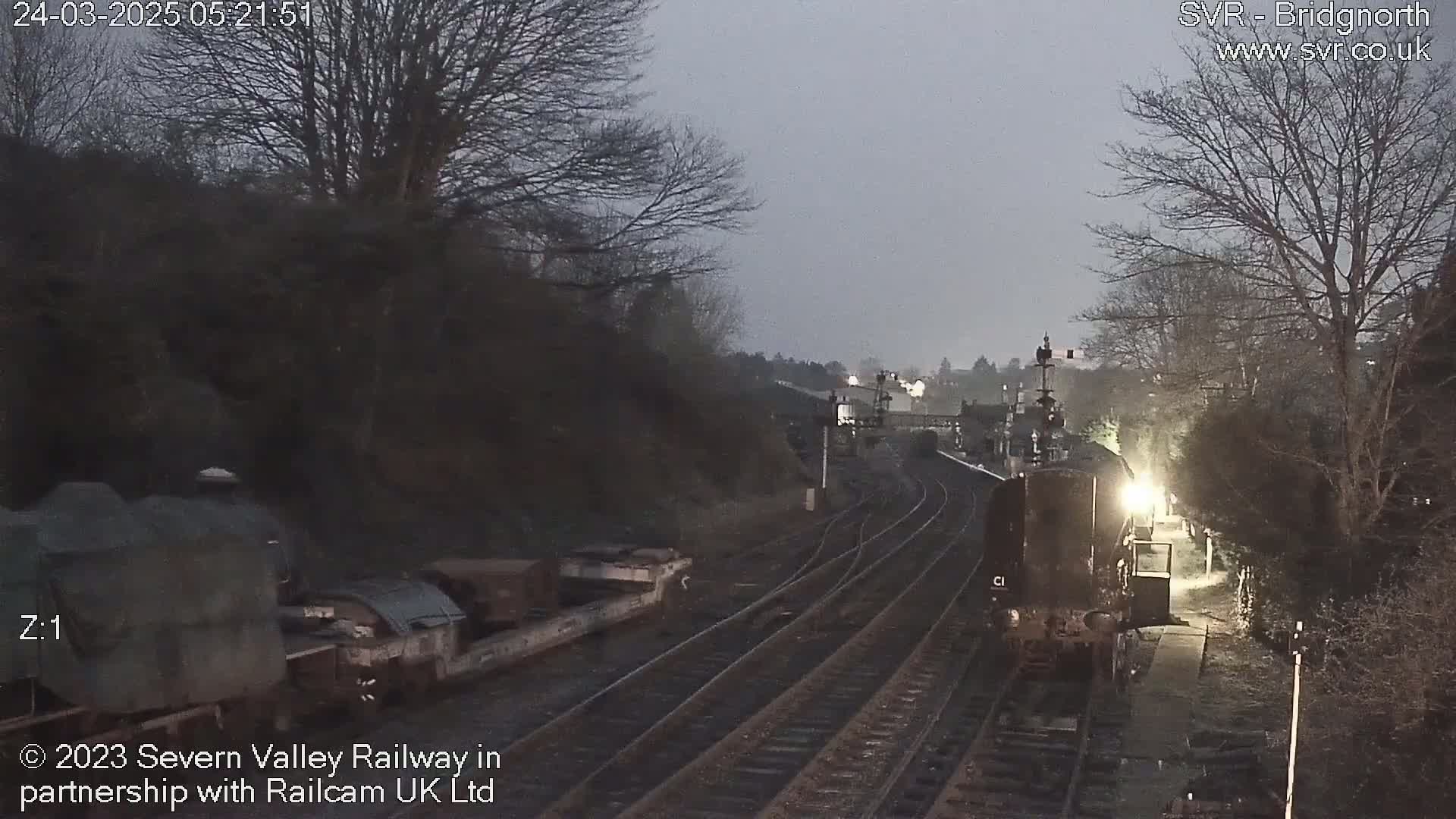 A diesel locomotive approaches a dimly lit train station at twilight, passing alongside a siding with stationary rail cars.