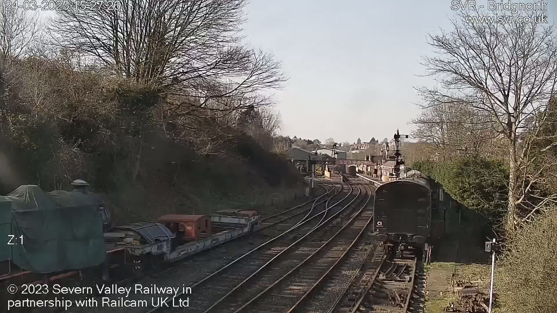 A dark-colored train moves along a railway track on a partly sunny day, passing by other railway cars and a wooded area.