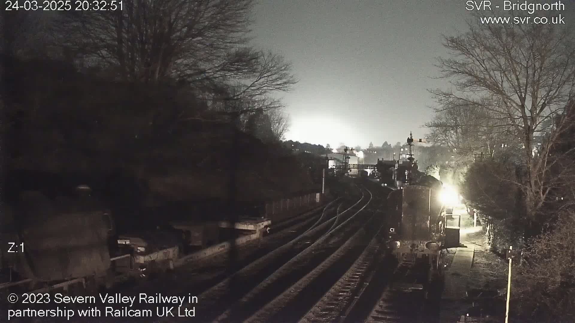 A nighttime view of train tracks splitting into multiple paths, surrounded by bare trees under a cloudy sky.