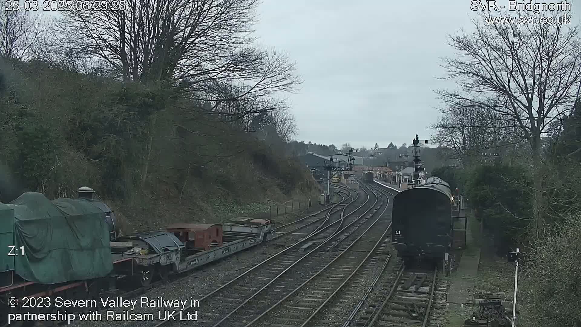 A dark-colored train is moving along train tracks in a railway yard on an overcast day, passing by other railway cars and buildings.