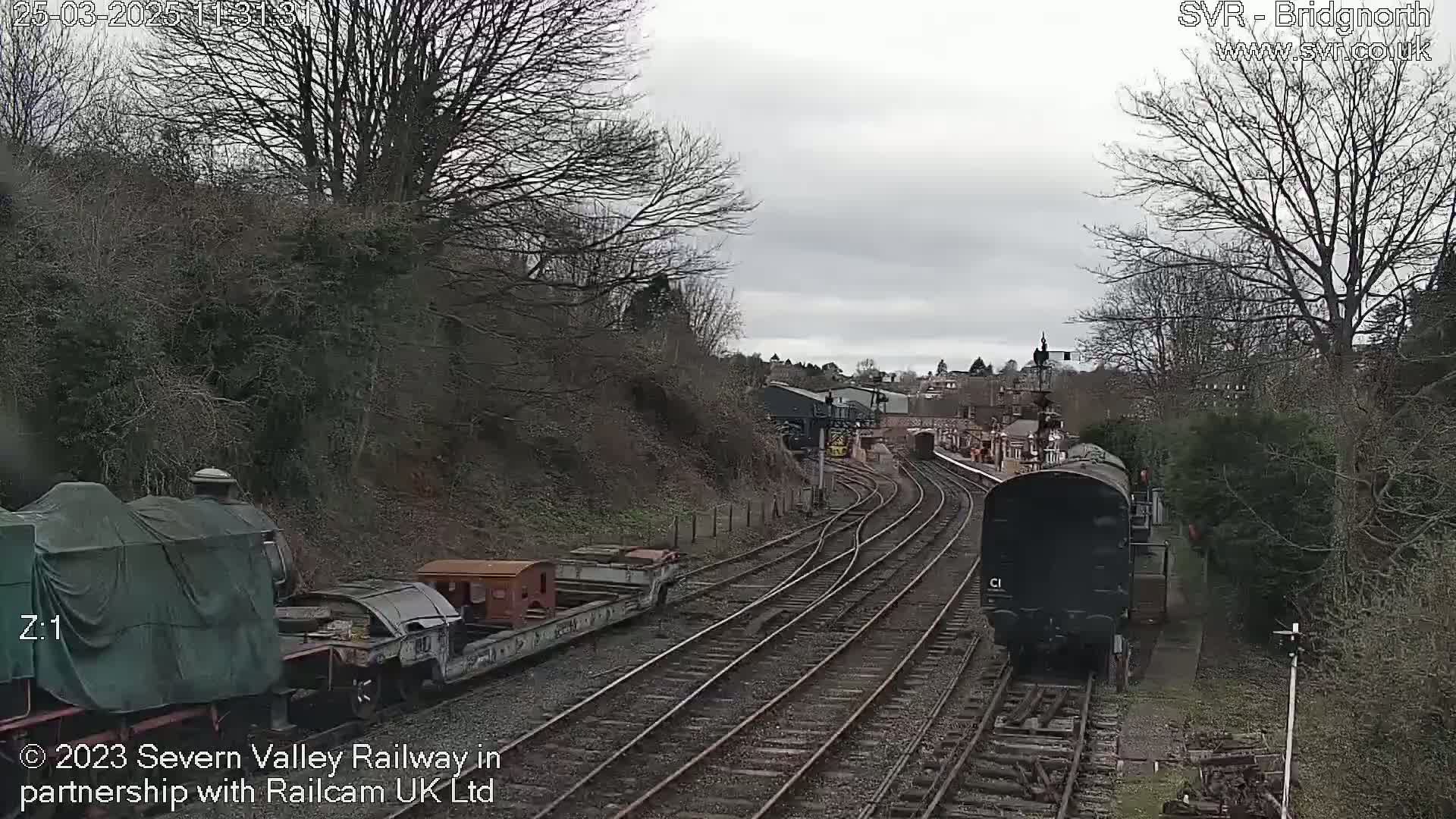 A dark-colored locomotive is traveling along a railway track through a barren landscape under an overcast sky.