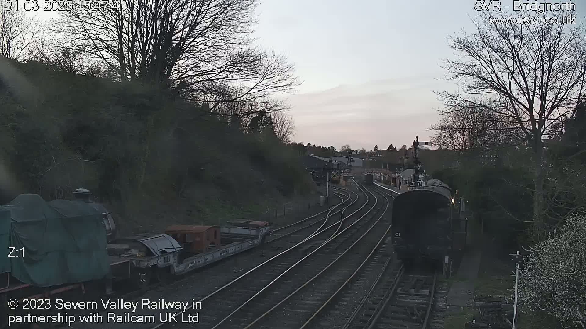 A steam train approaches a railway station under a twilight sky, passing several railway cars parked on a side track.