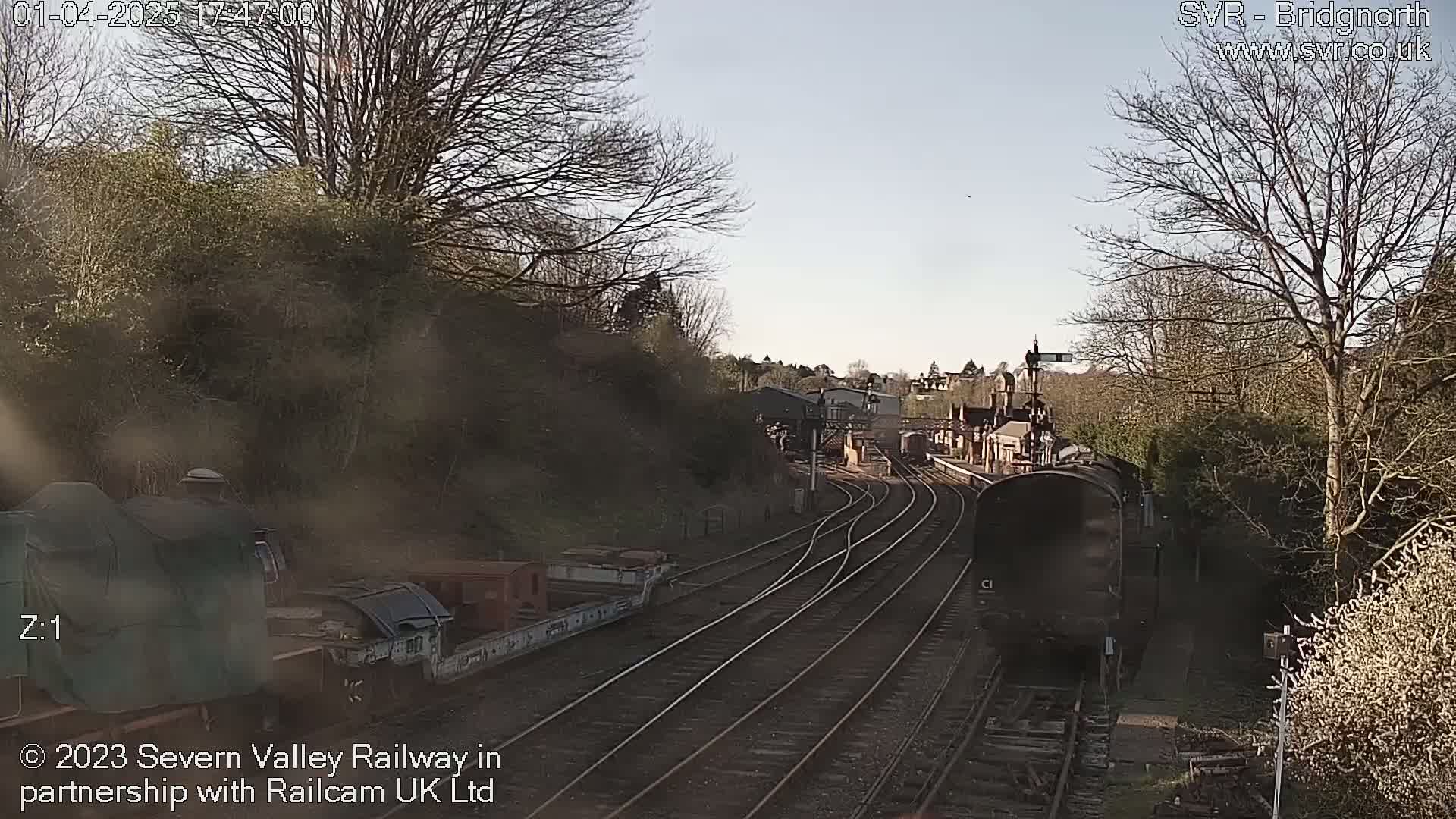 A steam train approaches a railway station on a sunny day, with bare trees lining the tracks and hillsides.