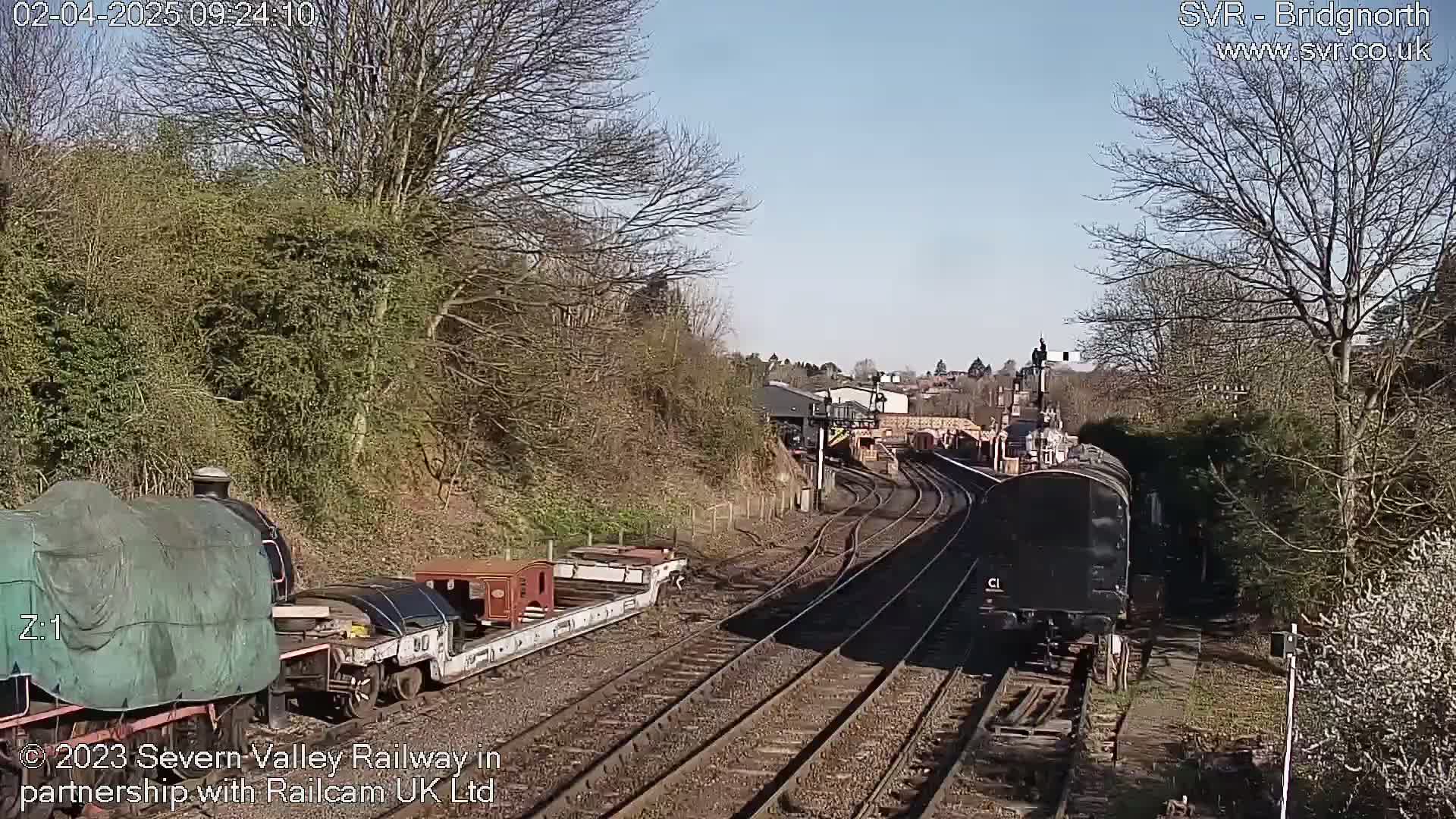 A sunny day shows railway tracks curving through a landscape of bare trees and shrubs, with several railcars and locomotives visible.