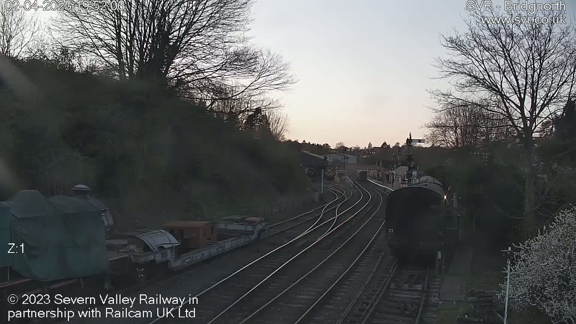 A steam train approaches a railway station at twilight, passing alongside several railway tracks and trees.
