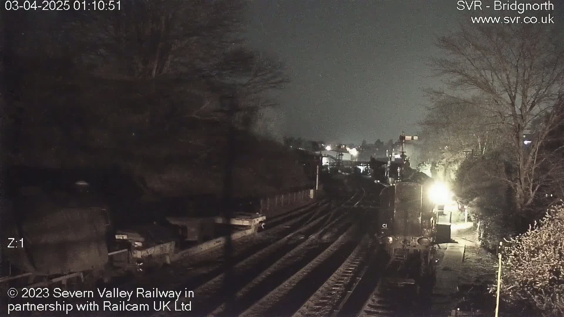 A nighttime view of train tracks and railway infrastructure, dimly lit under a dark sky.