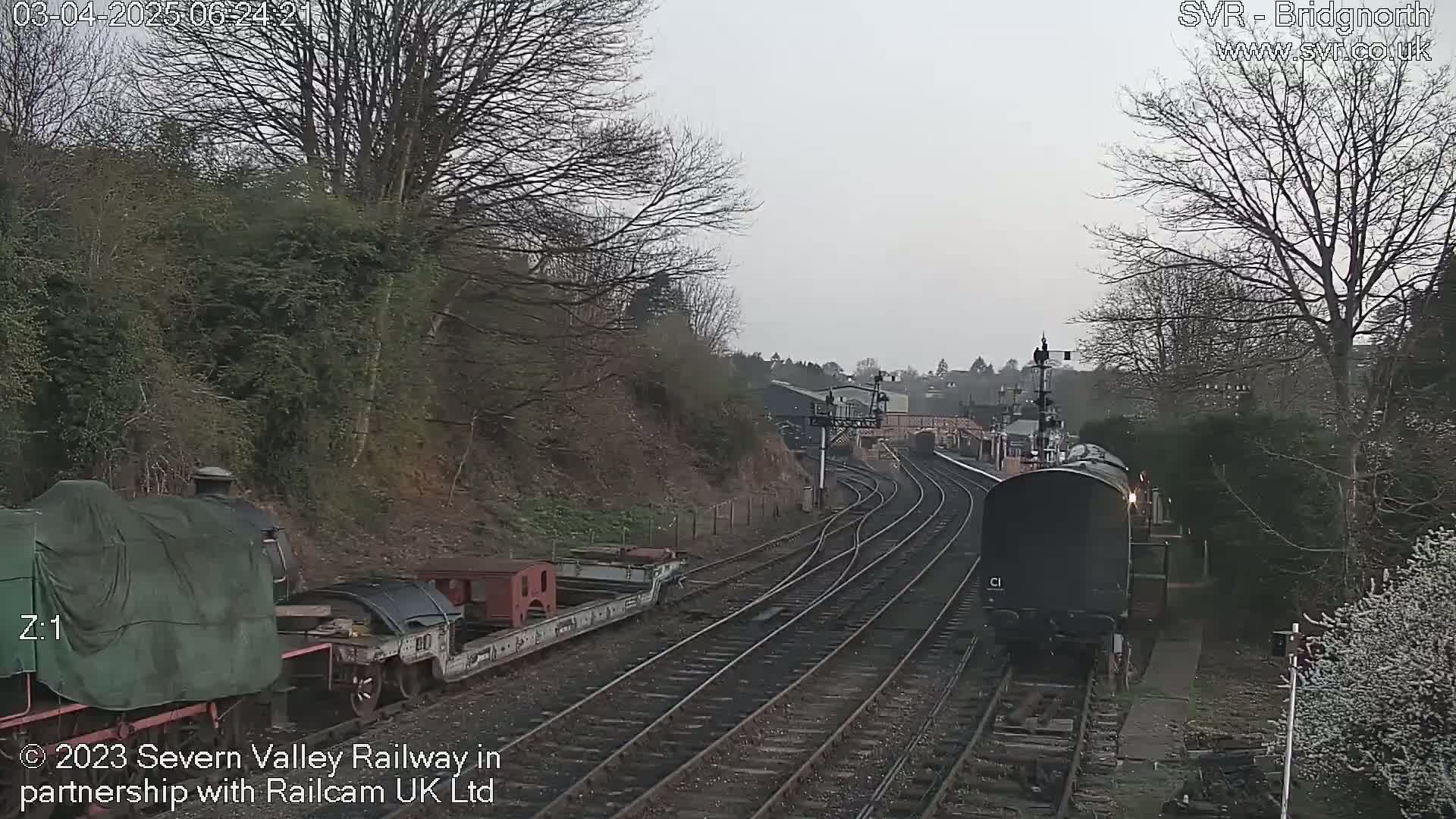A dark-colored train moves along a railway track on an overcast day, passing a stationary train partially covered by a tarp.