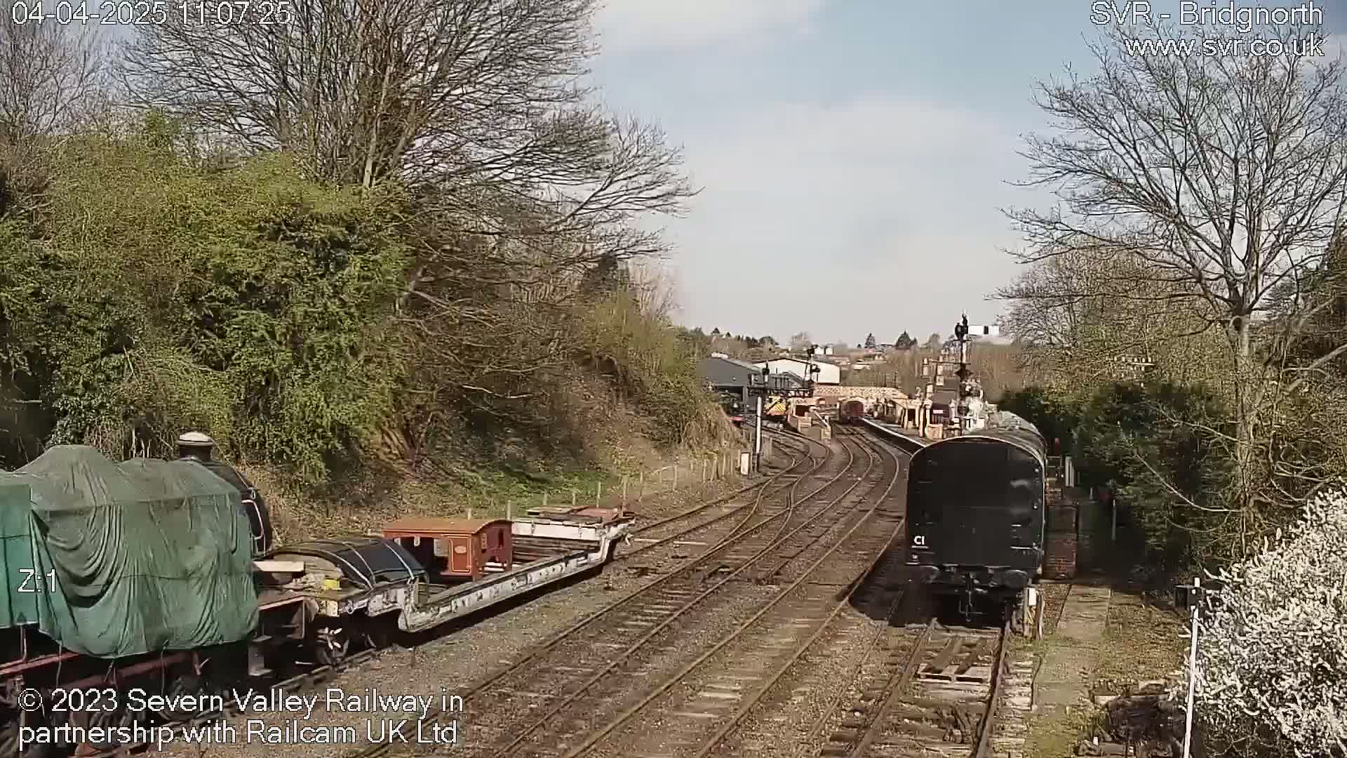 A railway scene on a partly sunny day shows multiple train cars, including a covered engine and a black diesel locomotive, on a track that curves through a landscape of bare trees and shrubbery.