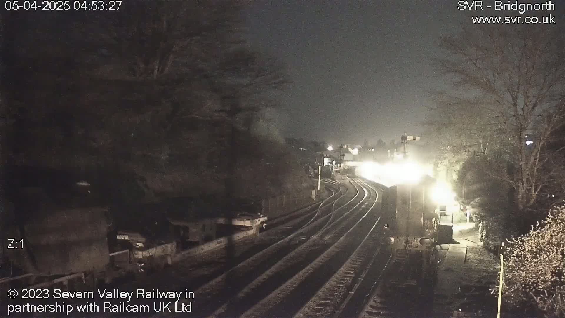 A brightly lit train approaches a dimly lit train station at night in snowy conditions.