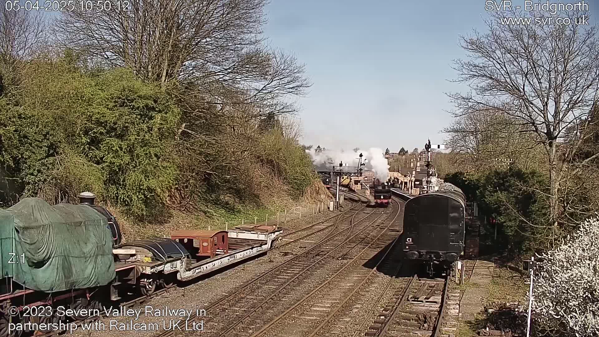 Two steam trains move along a railway track on a sunny day, with one train passing a station and the other parked nearby.