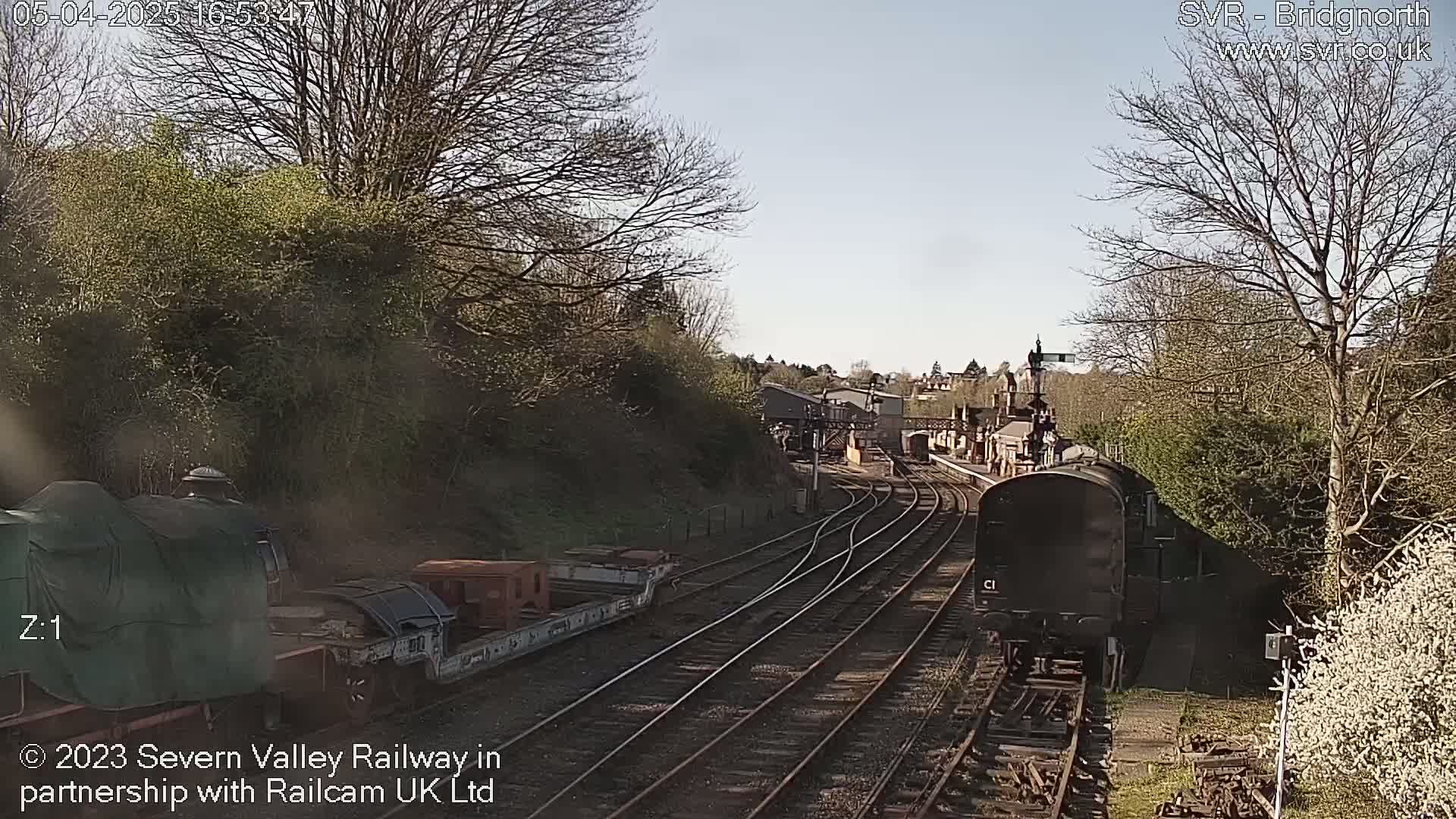 Two trains are visible on a railway track, one covered and stationary, and another dark-colored one moving, under a sunny sky with bare trees and budding vegetation.