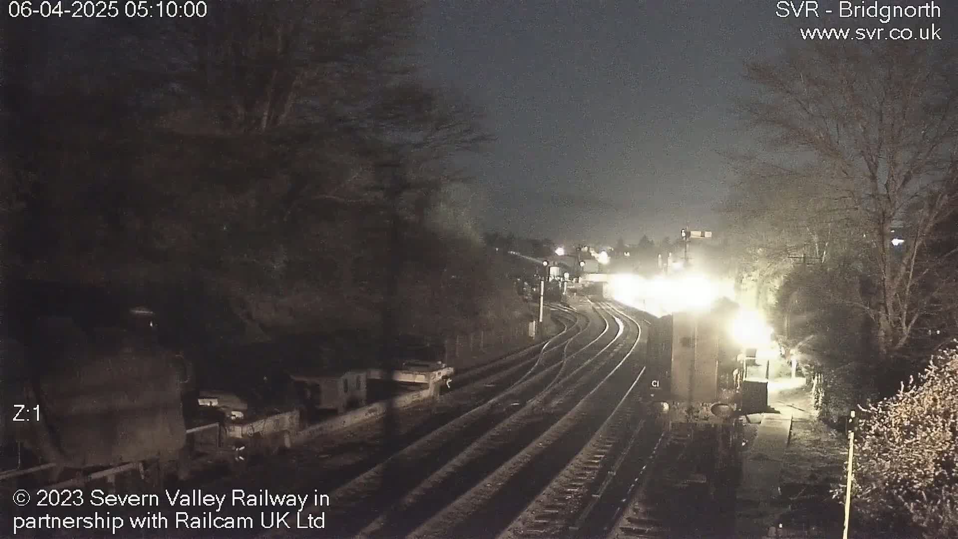A nighttime view of a train station with train tracks curving into the distance, illuminated by lights, surrounded by dark trees under a dark sky.