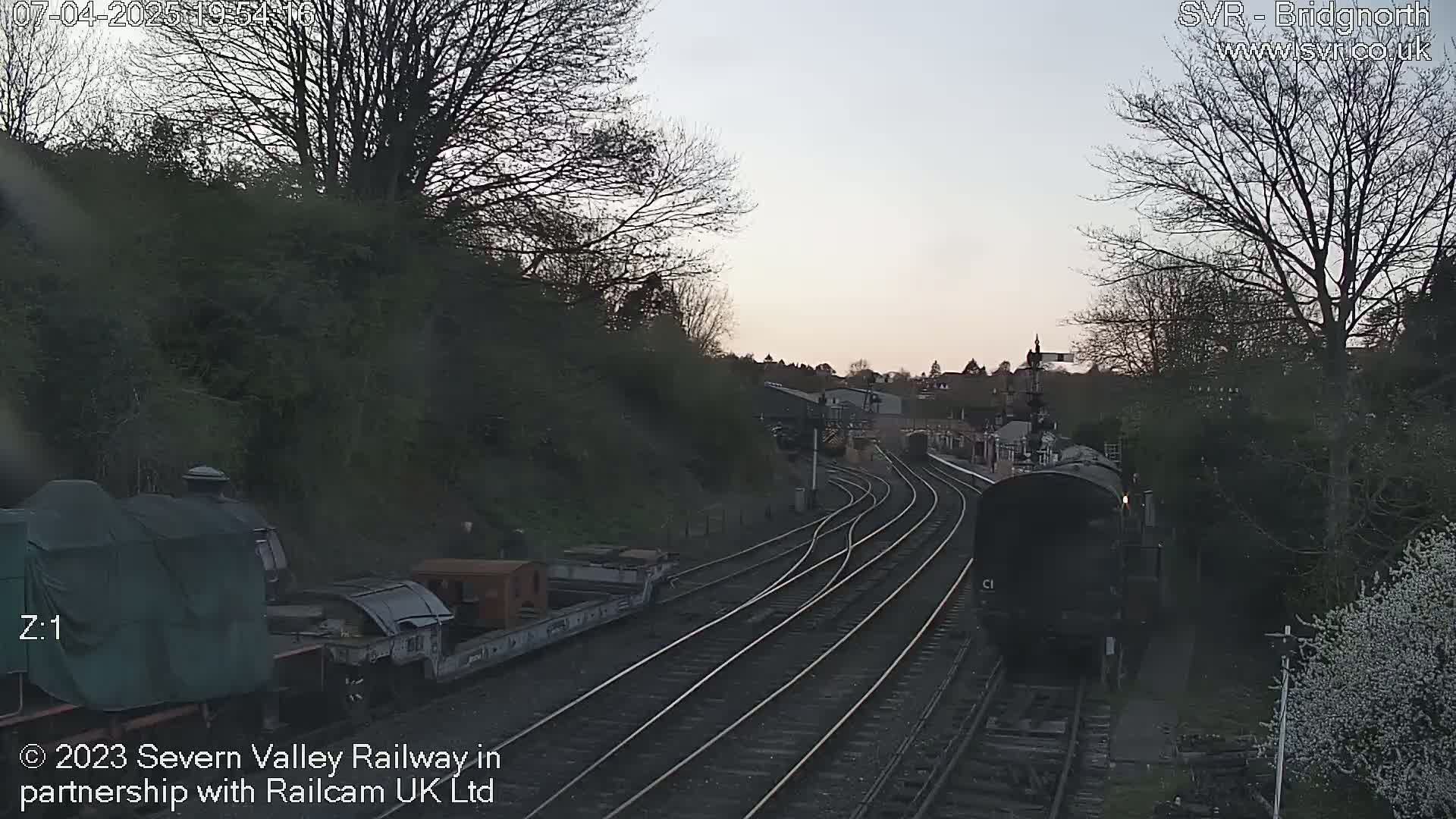 A train approaches a railway junction at dusk, passing alongside a stationary train and several railway sidings.
