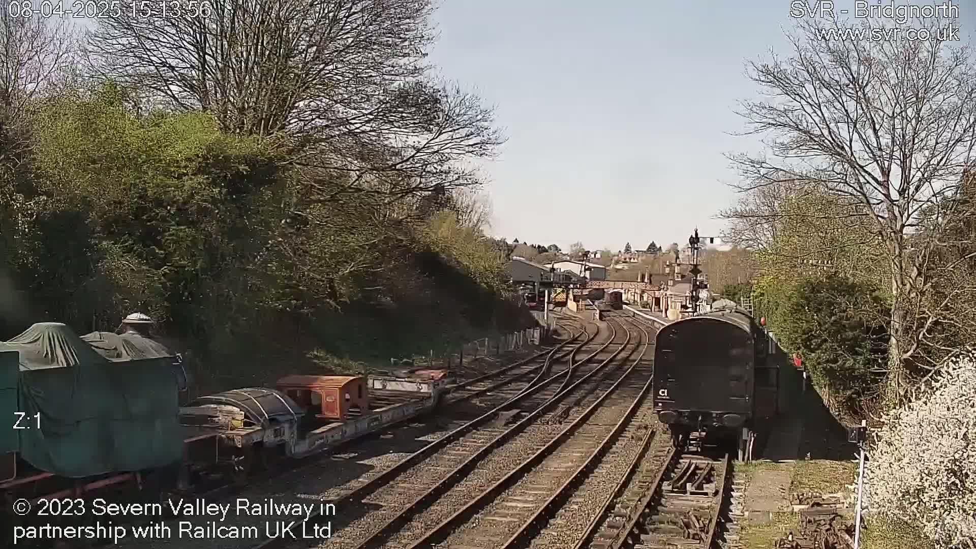 A sunny day at a railway station shows a black train car moving along tracks, with other rail cars and greenery visible.